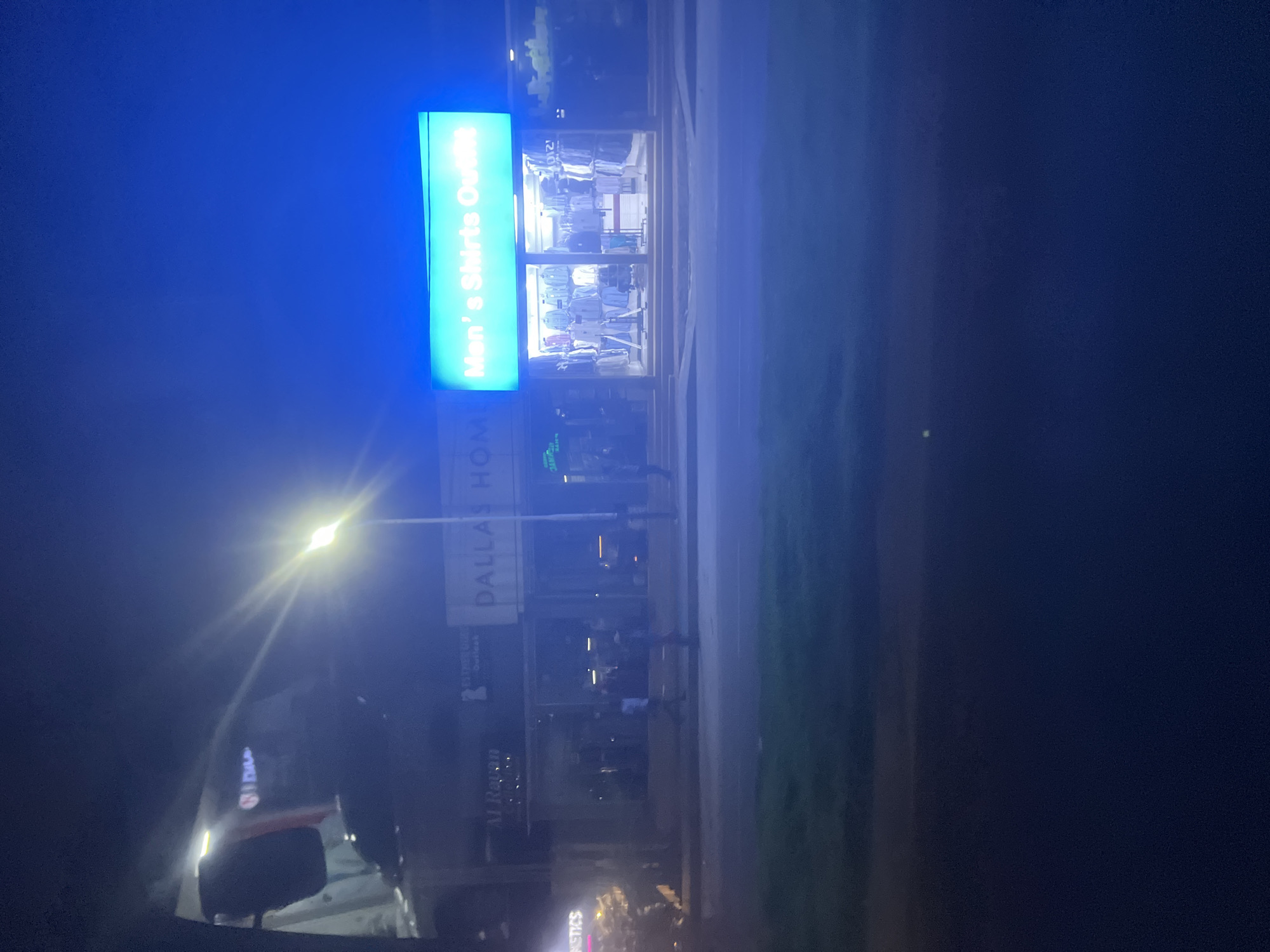 At night, the storefront for "Men's Shirts Outfit" under a blue signboard is lit up. Below this is "DALLAS HOME", with a sign below that too faint to read. There is an adjacent storefront below "Dallas Home" that is also too dark to read. A few individuals are seen in front of the store. A street runs along the right edge of the frame, with greenery beyond. Cars are parked along the street. The photograph seems to be taken from within a vehicle, looking out towards the storefront. The bright light of a streetlight is visible further down the street. The location is not readily identifiable as being in Dar es-Salaam, Tanzania.
