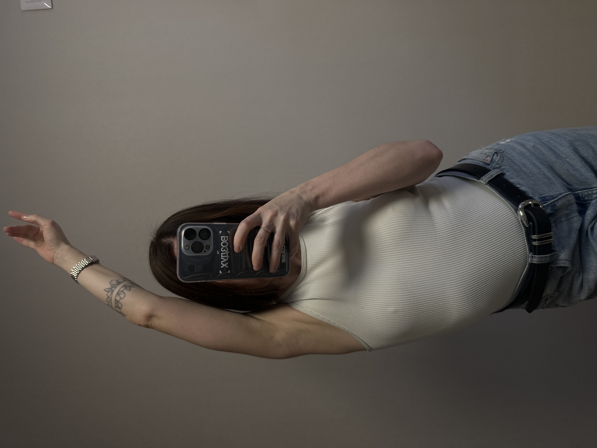 A person is taking a selfie in front of a plain, neutral-colored wall. The person is wearing a light-colored, ribbed tank top and light blue jeans with a black belt. A black cell phone case with the word "ВОЗДУХ" (AIR) written on it is visible. The person has a tattoo on their left forearm and a watch on their left wrist. There is a light switch on the wall in the upper left corner of the frame. The location cannot be determined from the image.