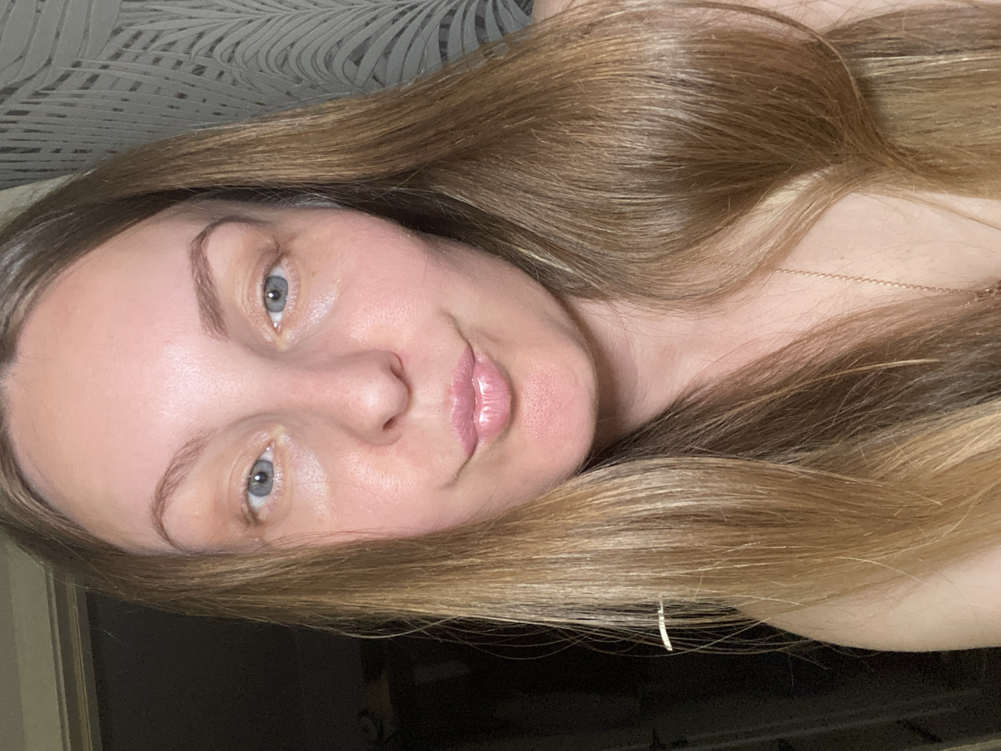 A woman with long, light brown hair and light blue eyes is looking at the camera. She has fair skin and light pink lips. She is wearing a delicate gold necklace. The background is blurry and shows a patterned wall.