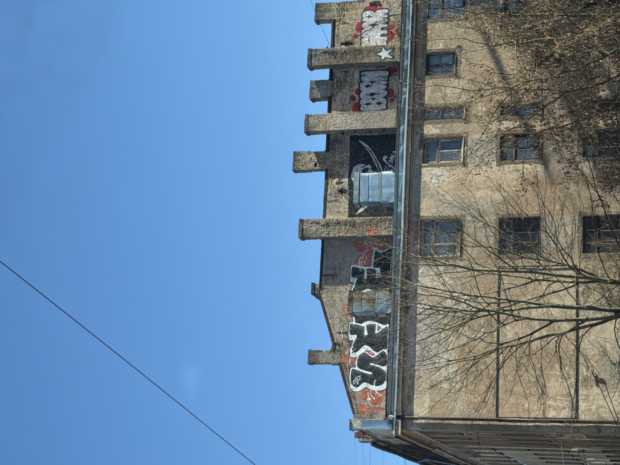 The image shows a low-angle view of a weathered building in Riga, Latvia, against a clear blue sky. The building is characterized by a concrete facade, with graffiti covering several sections. The building's structure includes protruding architectural elements reminiscent of a battlement. Windows are visible along the building's right side, partially obscured by leafless tree branches. A power line cuts across the bottom left of the sky.