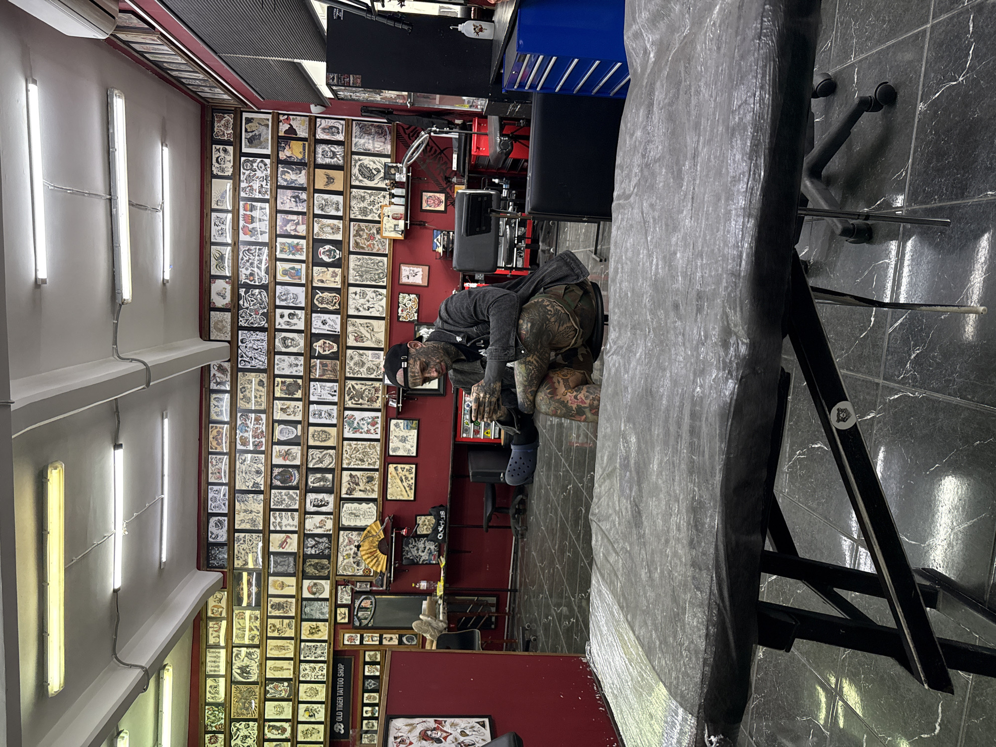 The image depicts the interior of a tattoo shop, likely in Moscow, Russia, given the context provided. The shop is lined with numerous framed tattoo flash art displayed on the walls. In the foreground, a tattoo table, covered with a grey plastic sheet, is visible. A man with extensive tattoos on his face and body is seated on a chair in the midground, looking at a mobile device. He is wearing a black baseball cap, a denim jacket, and camouflage pants. The floor is tiled with a dark, marbled pattern. Various pieces of tattoo equipment, including chairs and a blue rolling tool chest, are scattered throughout the space. The lighting appears to be from fluorescent ceiling fixtures.