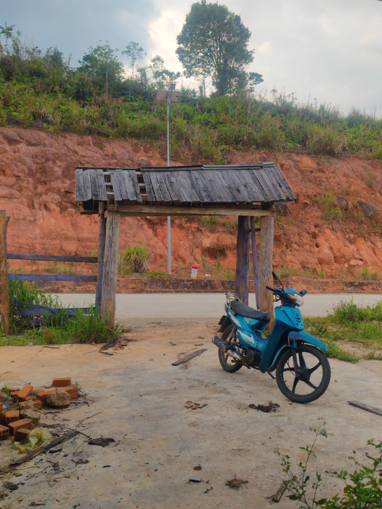The image depicts a rural roadside scene in Đam Rông 4, Vietnam, under an overcast sky. In the foreground, a turquoise blue underbone motorcycle is parked on a concrete patch next to a rustic wooden archway. The archway is constructed from rough wooden posts and planks, its roof appearing weathered and partially broken.
Behind the archway, a paved road stretches, flanked by a steep, red-orange earth hillside covered densely with green grass, bushes, and trees. A tall, slender pole topped with a solar panel stands behind the archway, slightly to the right, indicating a solar-powered light or camera. A smaller red and white marker pole is visible by the roadside, displaying the text "H9" and partially obscured letters, possibly "QT," below it.
To the left of the archway, a wooden fence or railing, painted partly blue, runs along the edge of the concrete area, with grass growing around its base. The ground in the immediate foreground is strewn with debris, including loose red bricks, scattered dry leaves and twigs, and some discarded plastic. No people are present, and the scene conveys a sense of quiet stillness in a natural yet partly developed environment. The diffused light suggests it could be an overcast day, late afternoon, or early morning.
