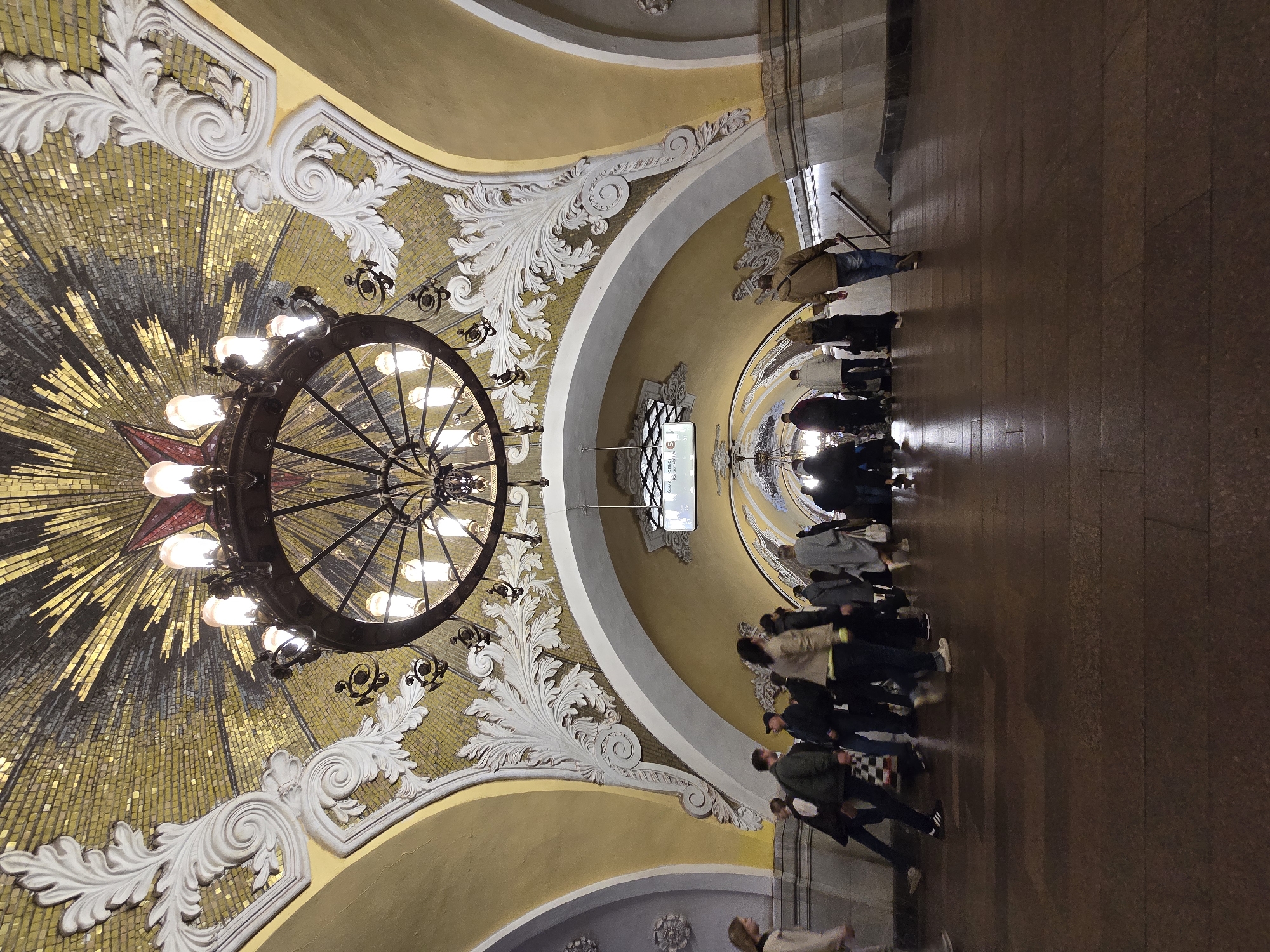 The image shows the interior of a Moscow Metro station in Russia. The perspective is angled upwards, emphasizing the architectural details. The station features an ornate, domed ceiling with decorative plasterwork, a golden mosaic pattern radiating from a central red star, and a large, circular chandelier. Below the ceiling, a passageway or platform is visible, with a group of people walking along it. The floor is made of polished brown stone. The overall ambiance of the scene is grand and historic.