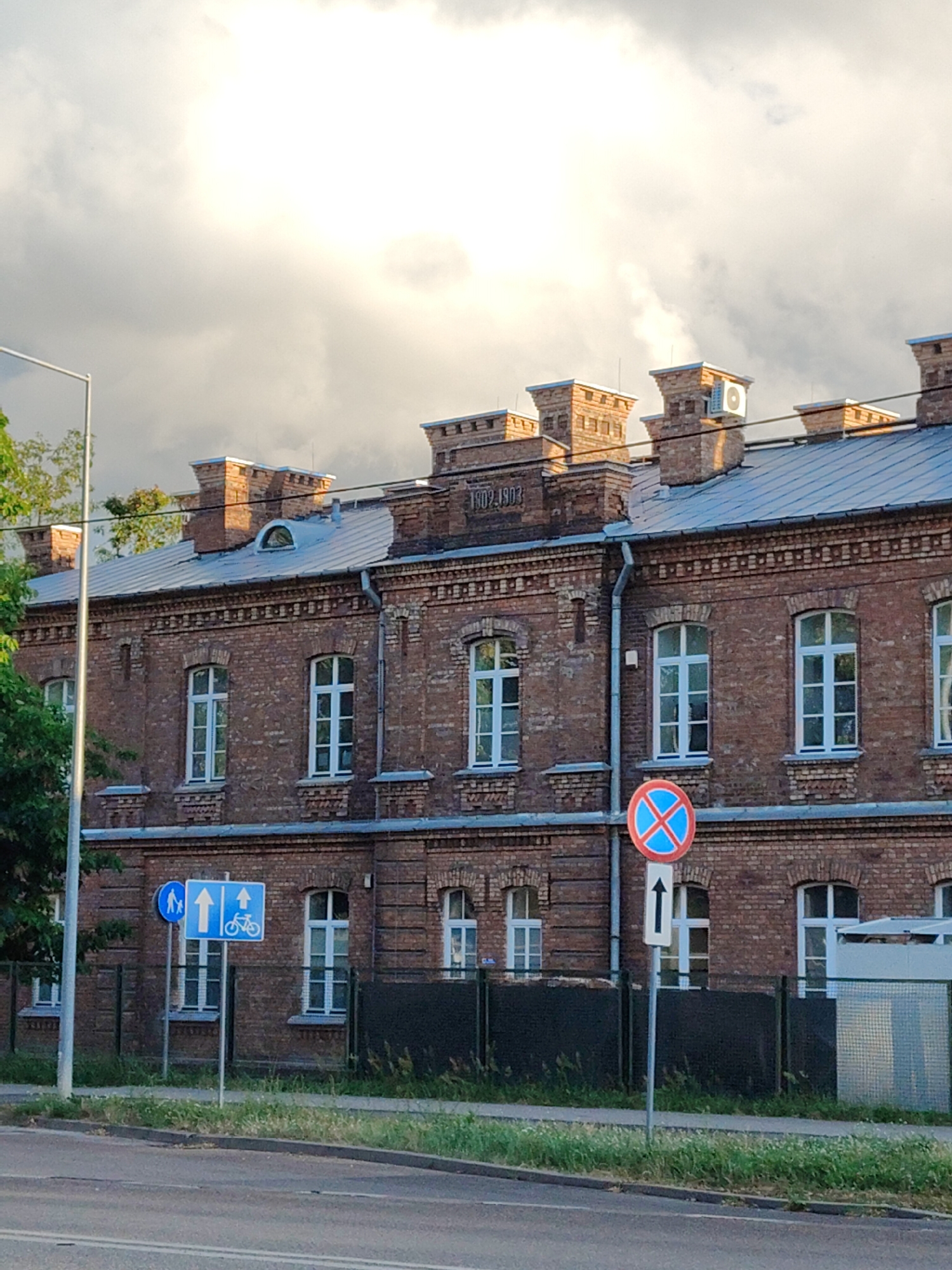 The image shows a brick building with multiple windows and a metal roof. Above the windows, there is text that reads "1802.1903." In front of the building, there are several traffic signs. One sign shows a person walking, a bicycle, and an arrow pointing upwards. Another sign shows a red circle with a blue cross inside, indicating no stopping. The building appears to be located on a street with grass and trees on the side. The sky is cloudy with sunlight breaking through. The overall scene suggests an urban environment in Nowy Dwór Mazowiecki, Poland.