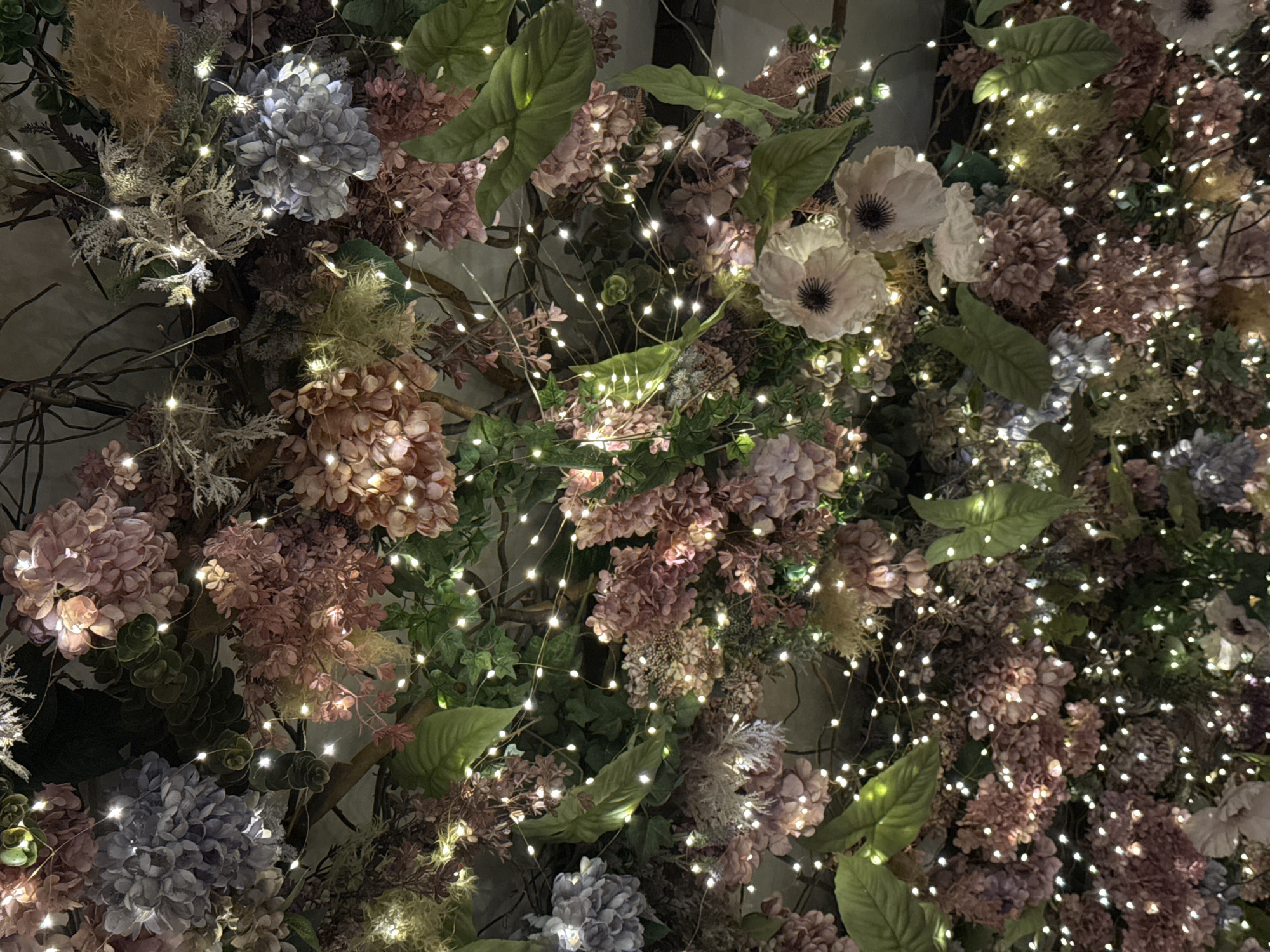 A dense floral arrangement covers a wall. The arrangement includes pink, blue, and white flowers interspersed with green leaves and brown branches. Twinkling fairy lights are woven throughout the floral arrangement, adding a soft glow. The background wall appears to be a light neutral color. The image provides no information to indicate a specific location in Sofia, Bulgaria.
