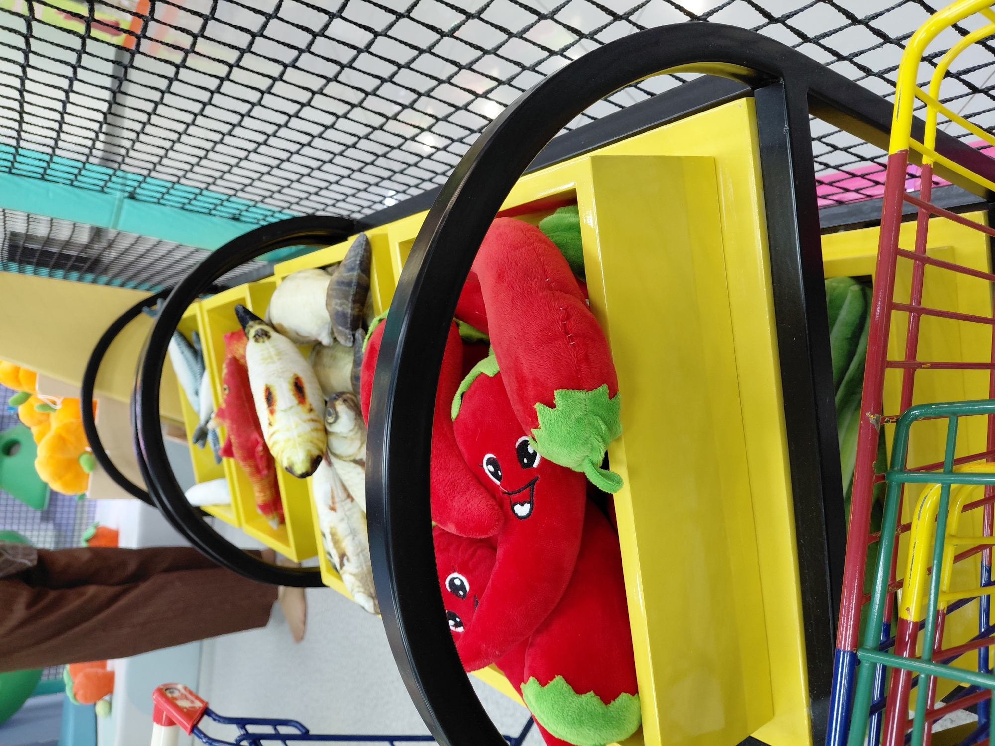 In an indoor children's play area, likely a play cafe, a yellow tiered display rack holds several plush toys. The top tier contains plush fish toys, some with realistic detailing. The second tier holds plush chili pepper toys with cartoonish faces. A black metal frame supports the yellow display rack. To the right of the display rack is a black safety net. In the foreground, a colorful metal shopping cart is visible. In the background, out of focus, a person is standing, and other play structures such as plush pumpkins and carrots can be seen. The scene is brightly lit. This image is not specific to Karaganda, Kazakhstan, and lacks any visual cues to pinpoint its location.
