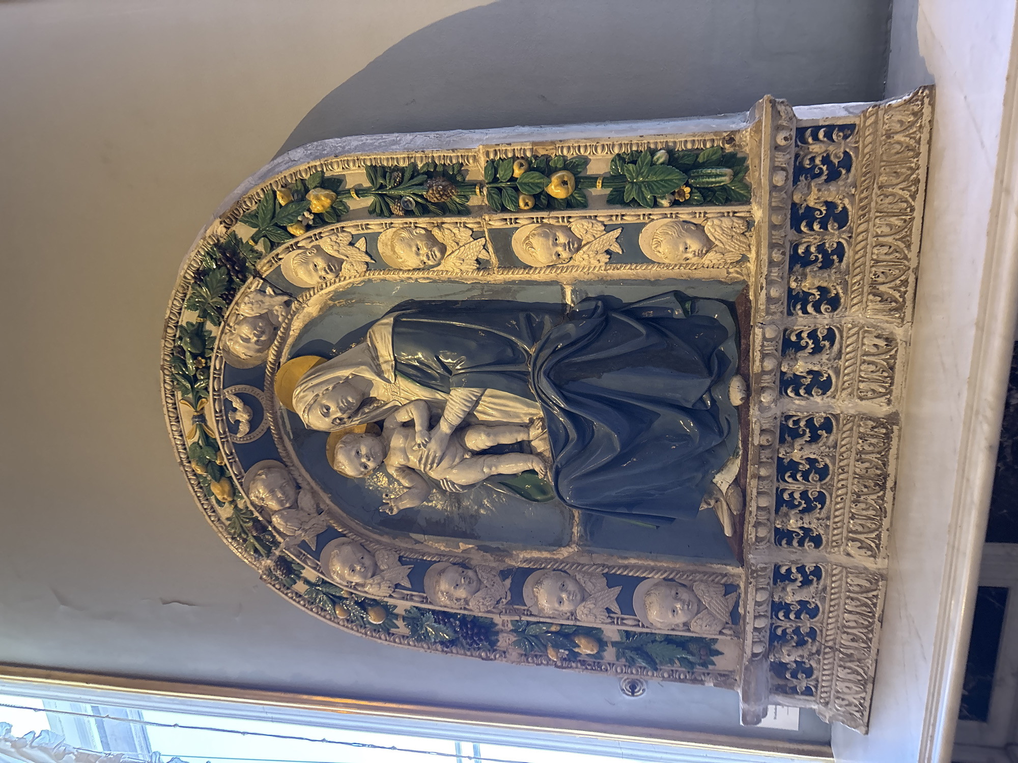 This image depicts a relief sculpture of the Madonna and Child, set within an ornate lunette. The central figures, the Virgin Mary and the infant Jesus, are rendered with soft, lifelike features. Mary is dressed in a deep blue robe, with her head tilted gently towards the child she cradles. The child is depicted nude, with a serene expression. They are positioned against a light blue background, suggesting a heavenly or serene setting.
The lunette itself is richly decorated. The inner border features a garland of green leaves, interspersed with yellow fruits and pine cones. Below this, a row of cherubic heads with small wings emerges from the background, looking outwards. The outer edges of the lunette are adorned with intricate relief work in white and gold, featuring scrolling patterns and what appear to be stylized animal figures in a dark blue inset.
The sculpture is mounted on a wall, and the lighting suggests it is indoors, possibly in a museum or religious building, given the subject matter. The overall impression is one of artistic craftsmanship and religious devotion. The style of the sculpture suggests it is Renaissance or Baroque in origin. There is no visible text in the image. The image is taken from a slightly elevated angle, looking upwards at the sculpture.