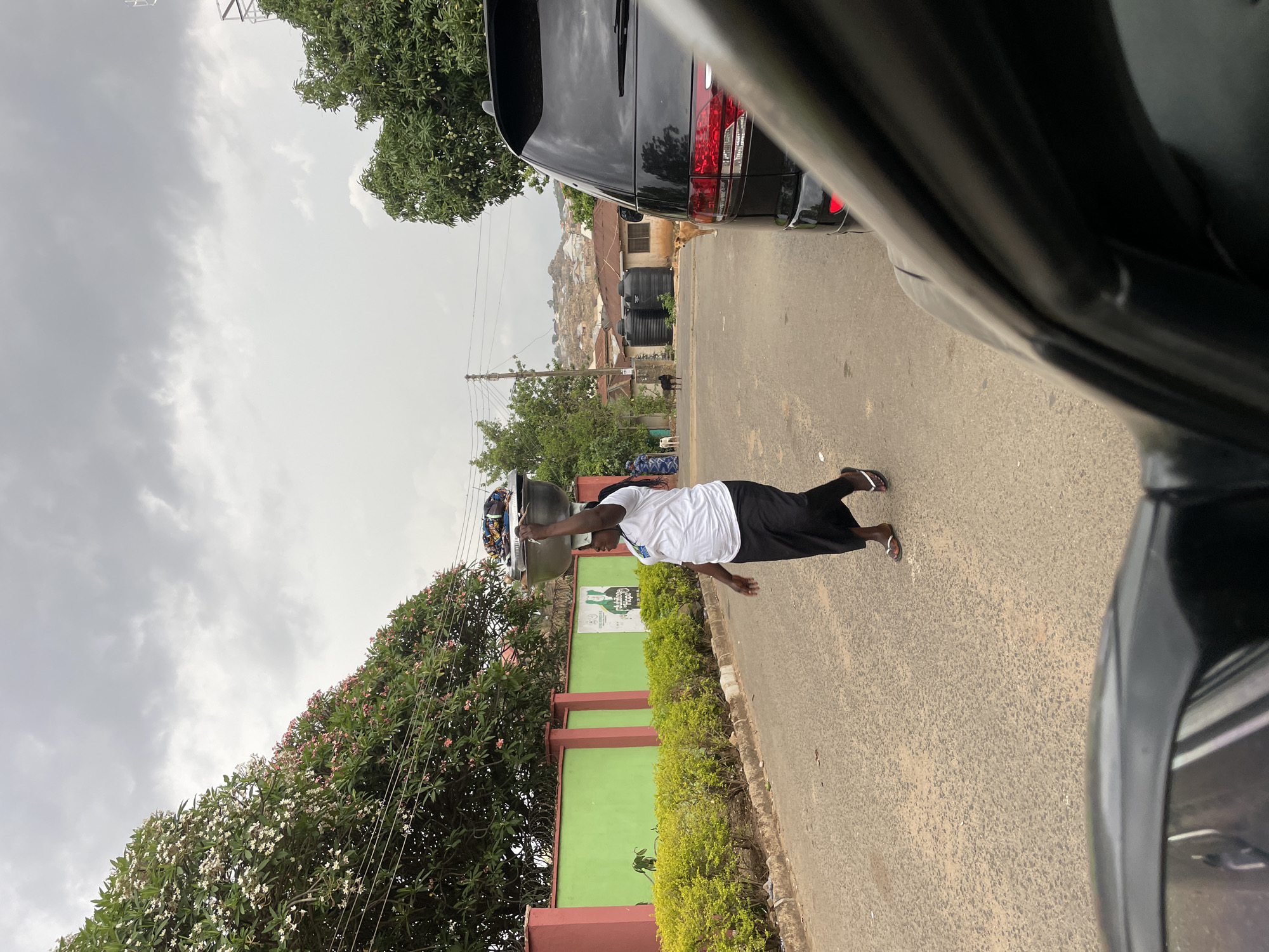 A person is carrying a large bowl on their head while walking along a street in Jos, Nigeria. The person is wearing a white t-shirt, a black skirt, and sandals. The street is paved and lined with trees and a green wall. In the background, there are houses built on a hillside. There is a black vehicle partially visible on the right side of the image. The sky is overcast.