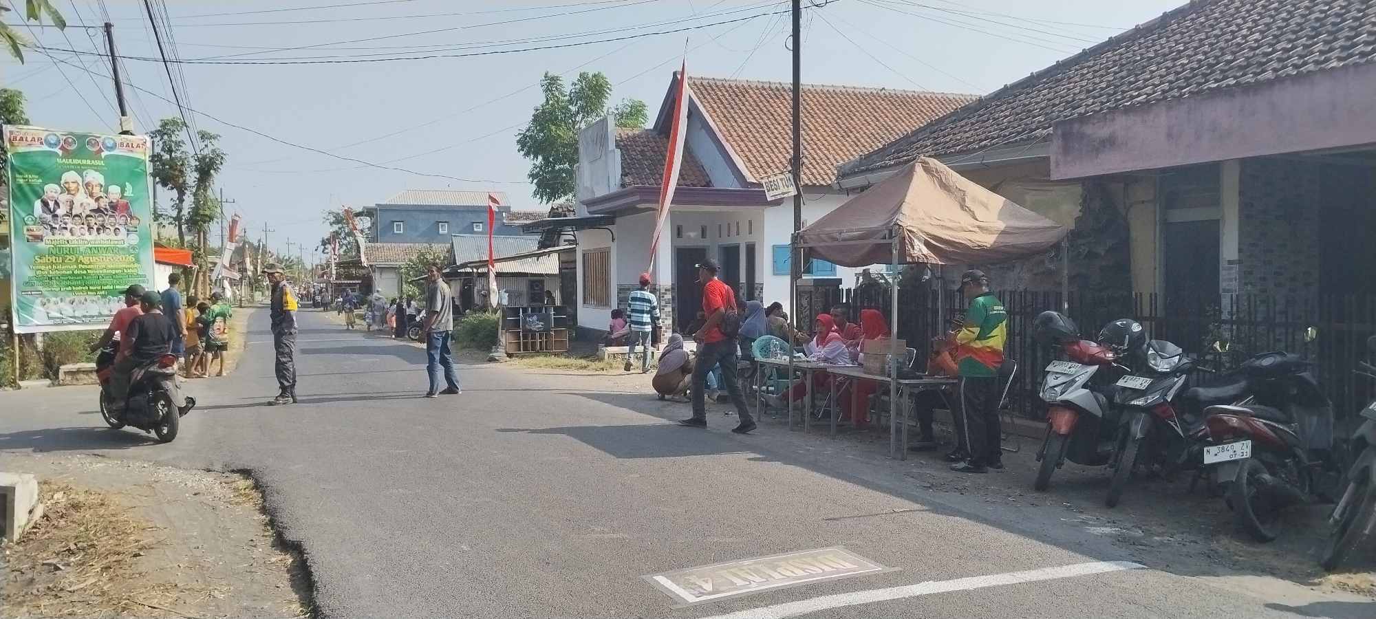 The image depicts a street scene in Yosowilangun Kidul, Indonesia. On the left side of the road, a large banner is displayed, advertising a religious event called "MAULIDURRASUL NURULANWAR" scheduled for Saturday, August 29, 2026. Several people are gathered near the banner, some on a motorcycle. A person in uniform is standing on the road, seemingly directing traffic or providing security.
The road itself is paved and appears to be a two-lane street. On the right side of the road, there are houses with traditional Indonesian architecture, some decorated with red and white Indonesian flags. A temporary tent is set up in front of one of the houses, with several people seated at tables, possibly for an event registration or a community gathering. Various motorcycles are parked along the side of the road. The environment is outdoors, under a clear sky, suggesting a daytime event. The overall scene suggests a community gathering or event taking place in a residential area.