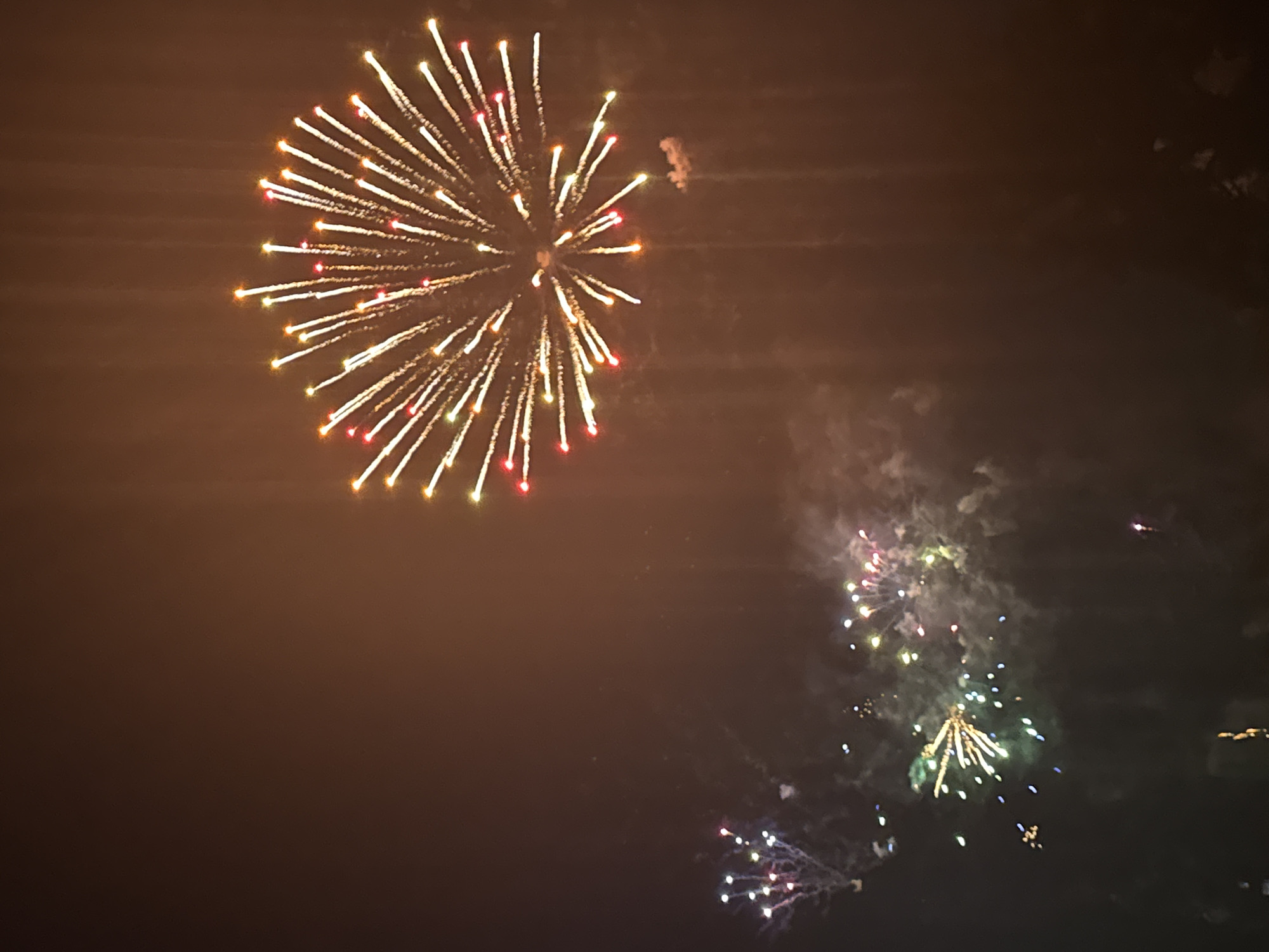 The image depicts a nighttime fireworks display. A large, bright firework explodes in the upper right quadrant, with numerous thin, radiating lines of light in shades of yellow, white, and red. Smaller fireworks are visible in the lower left and middle of the frame, characterized by clusters of colored lights in white, green, and pink. The background is a dark, hazy sky, suggesting a smoky atmosphere from the fireworks. The overall impression is of a festive celebration. No specific landmarks or elements that would definitively place this event in Lille, France, are visible within the frame.