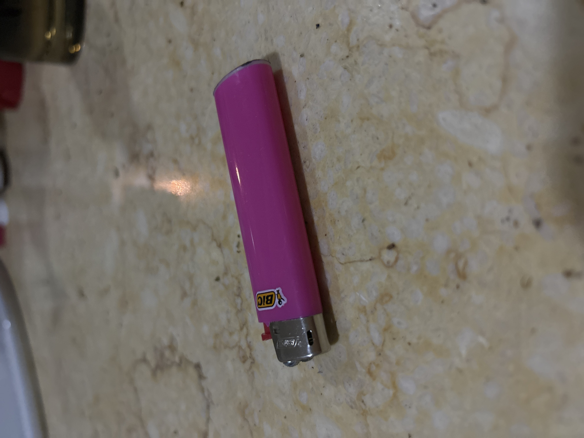 A pink Bic lighter rests on a light beige countertop. The lighter is in the center of the frame and positioned at an angle. The countertop has a mottled pattern, with various shades of beige, tan, and brown. Part of a jar and a white basin are visible in the left corner. The location is not discernible based on the provided image.