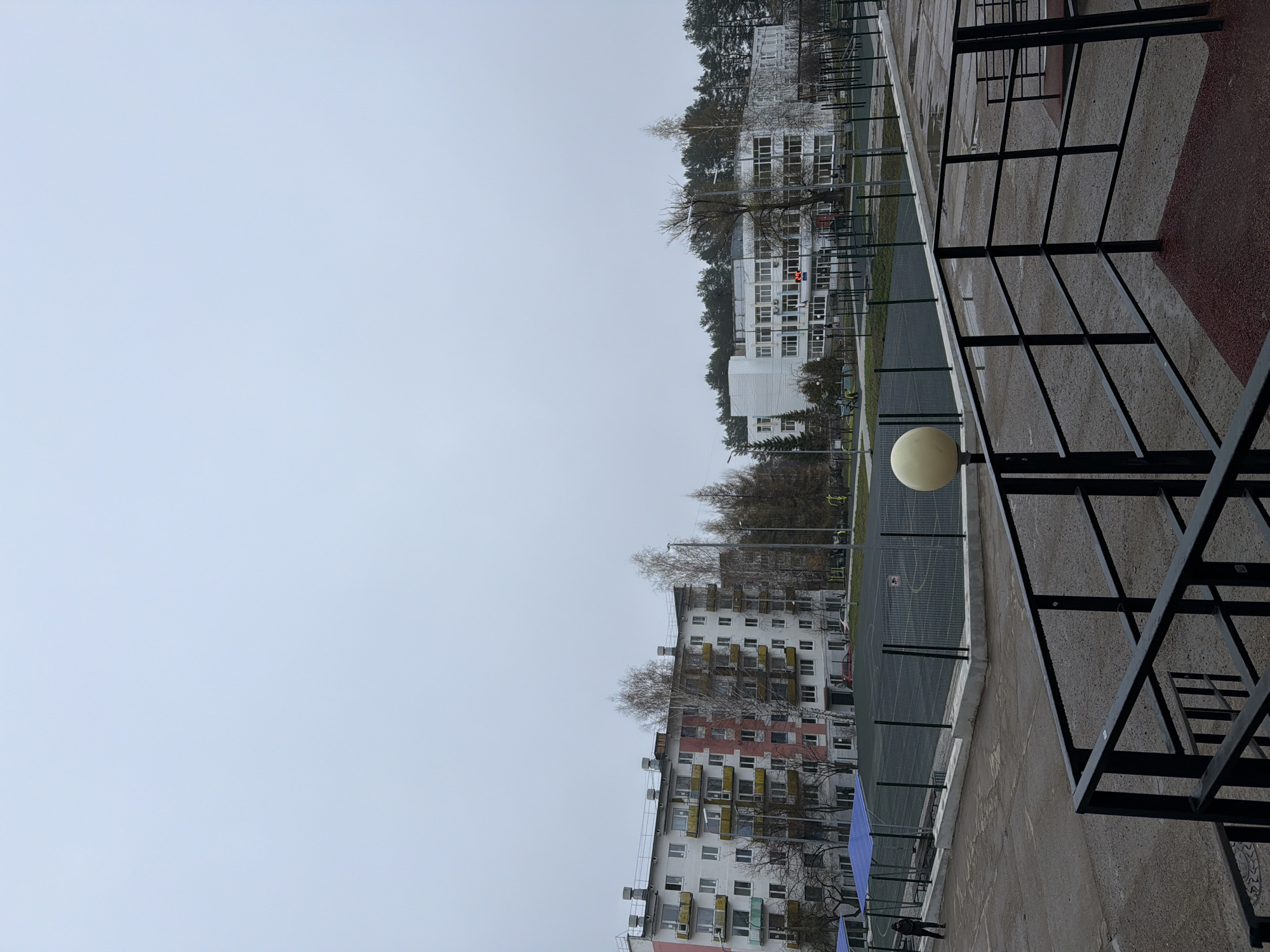 This image, taken in Chaikovsky, Russia, depicts an outdoor urban setting under an overcast sky. The scene is bisected by a long, grey horizon, separating a vast expanse of light grey sky from the lower landscape.
In the middle ground, two multi-story buildings are prominent. To the left (when oriented correctly), a residential apartment building features a mix of white, red/pink, and yellow sections, with numerous balconies, some enclosed. On the right, a large white building, possibly a school or public facility, stands with many windows. A digital display on this building shows "13:20."
Between these buildings lies a fenced sports court, likely for basketball or football, with visible court markings and a faint basketball hoop structure near the white building. Bare deciduous trees are scattered throughout the scene, particularly behind and between the buildings, suggesting a cool season like late autumn, winter, or early spring. A small, dark vehicle is parked near the white building.
The foreground on the right side of the image features a robust black metal railing or structure, possibly part of a fence or outdoor gym equipment. A spherical, cream-colored outdoor light fixture is positioned in the lower right. The immediate foreground consists of a concrete or paved area, with a section of red-surfaced ground, possibly a running track, visible on the far right. A blue tarp or covering is visible near the base of the apartment building on the left. A solitary, indistinct person is faintly visible in the lower left, near the apartment block. The overall atmosphere is quiet and still, with no specific activity clearly taking place.