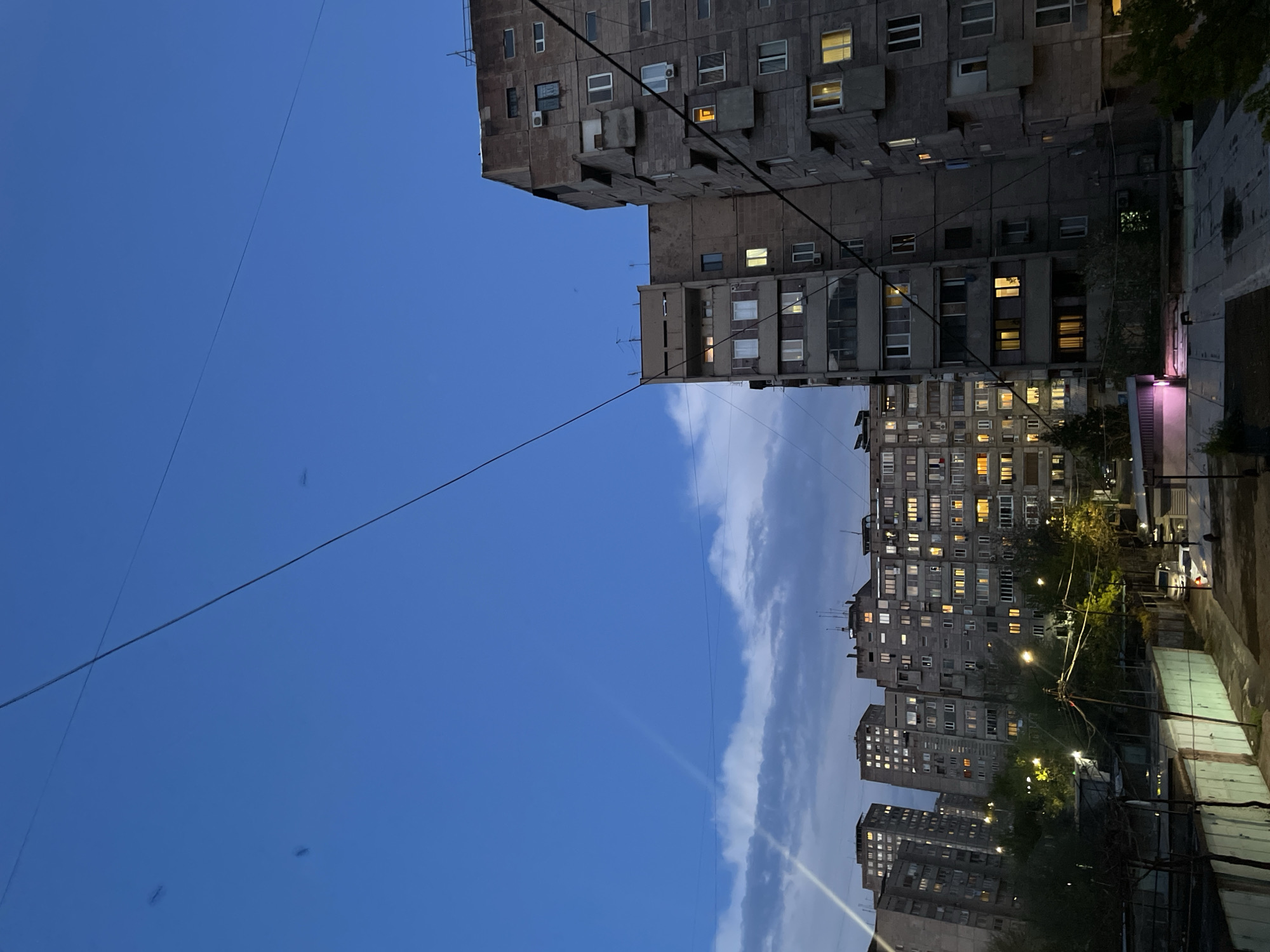The image captures an urban landscape at twilight, likely in Yerevan, Armenia. The scene is dominated by several multi-story residential buildings, characteristic of post-Soviet architecture, made of grey concrete with numerous windows. Many of the windows in the buildings are illuminated with warm, golden light, indicating human presence and the time of day.
The foreground and midground feature multiple large, monolithic apartment blocks that extend vertically across the frame. These buildings have a somewhat stark and functional appearance, with various extensions and balconies visible. The building in the center-right is particularly prominent, showing a dense array of windows, many lit. To the right, another building or structure displays a distinct pinkish-purple light emanating from a lower level. Streetlights are also visible, casting bright cones of light on the ground and nearby foliage.
The upper-left portion of the image is dominated by a deep blue sky, transitioning from a lighter hue near the horizon to a darker shade overhead, typical of dusk or early evening. A long, thin cloud formation stretches horizontally across the middle of the sky, adding texture. Several thin utility wires crisscross the sky, connecting various points and disappearing into the distance.
No specific human activity is discernible, but the illuminated windows convey a sense of bustling urban life within the buildings. There is no visible text in the image. The overall atmosphere is quiet and observant, capturing a moment in a residential area as day transitions to night.