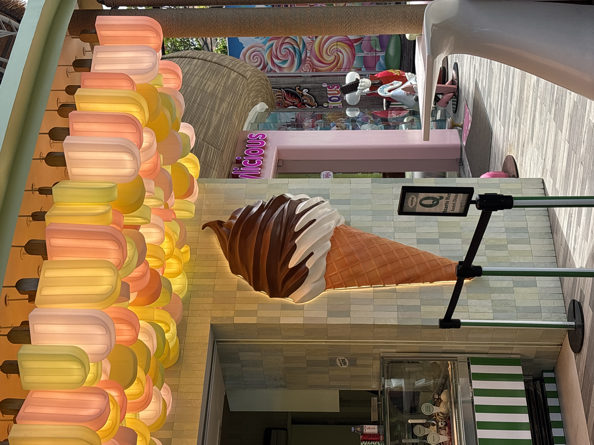 The image depicts an ice cream shop exterior, likely located in Denpasar, Indonesia. The shop's facade features an arrangement of popsicle-shaped lights in pastel hues on one side, and a large, illuminated ice cream cone mounted above the entrance. The shop is identified by a sign that reads "olicious" above the door.
Above, a candy-themed mural with lollipops is seen. The entrance has a glass display. In the background, a white chair is visible. The image is taken at ground level, capturing the storefront and surrounding elements. There are signs for the entrance line to Antri Disini.