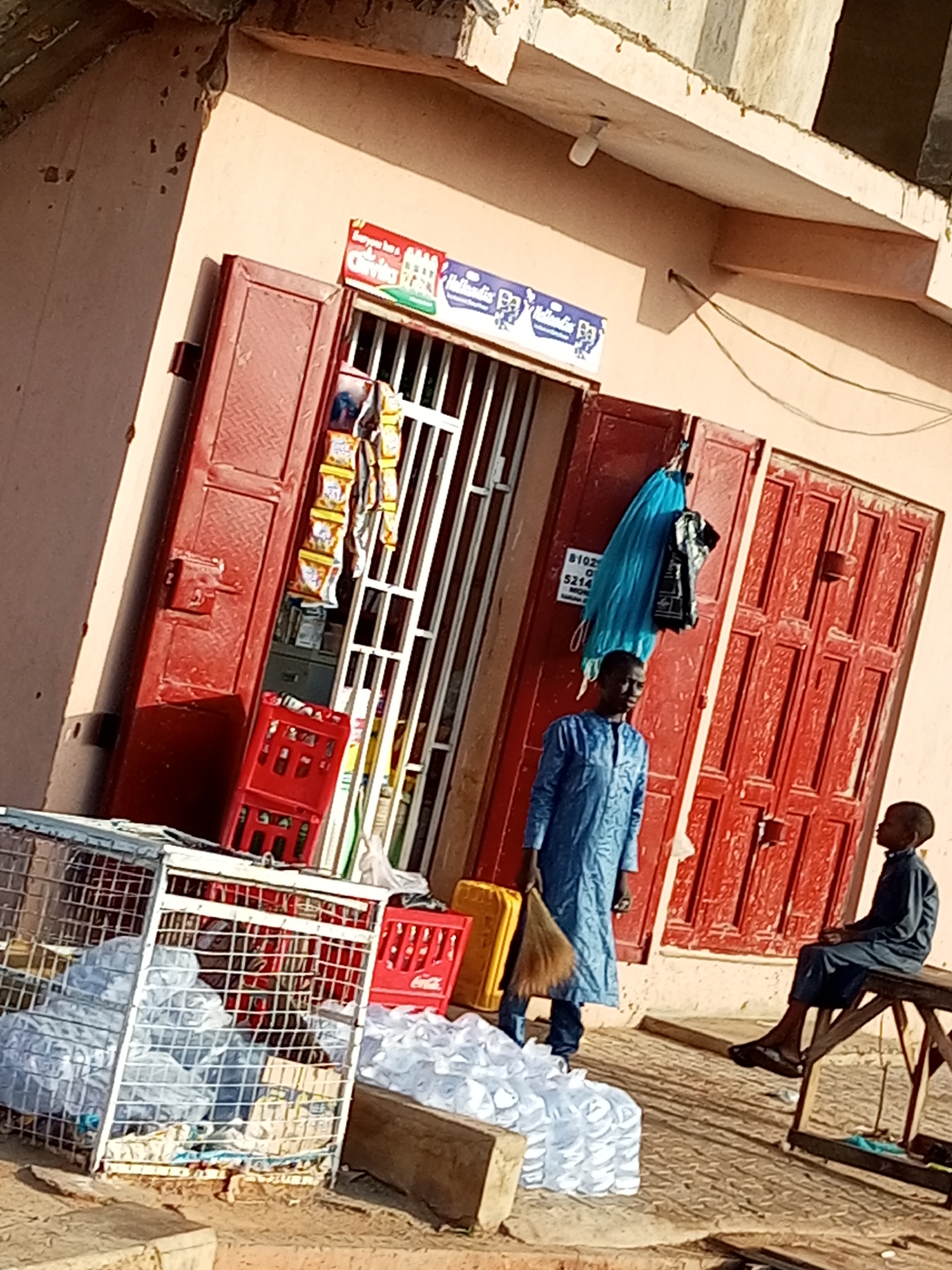 In the city of Gombe, Nigeria, a shop with red double doors is open. A man in a blue garment stands to the left of the entrance, holding a broom. A young boy in a dark garment sits on a wooden bench to the right of the entrance. In front of the shop, there are several crates of drinks and cages filled with water bottles. The scene appears to be a marketplace or a street-side vendor.