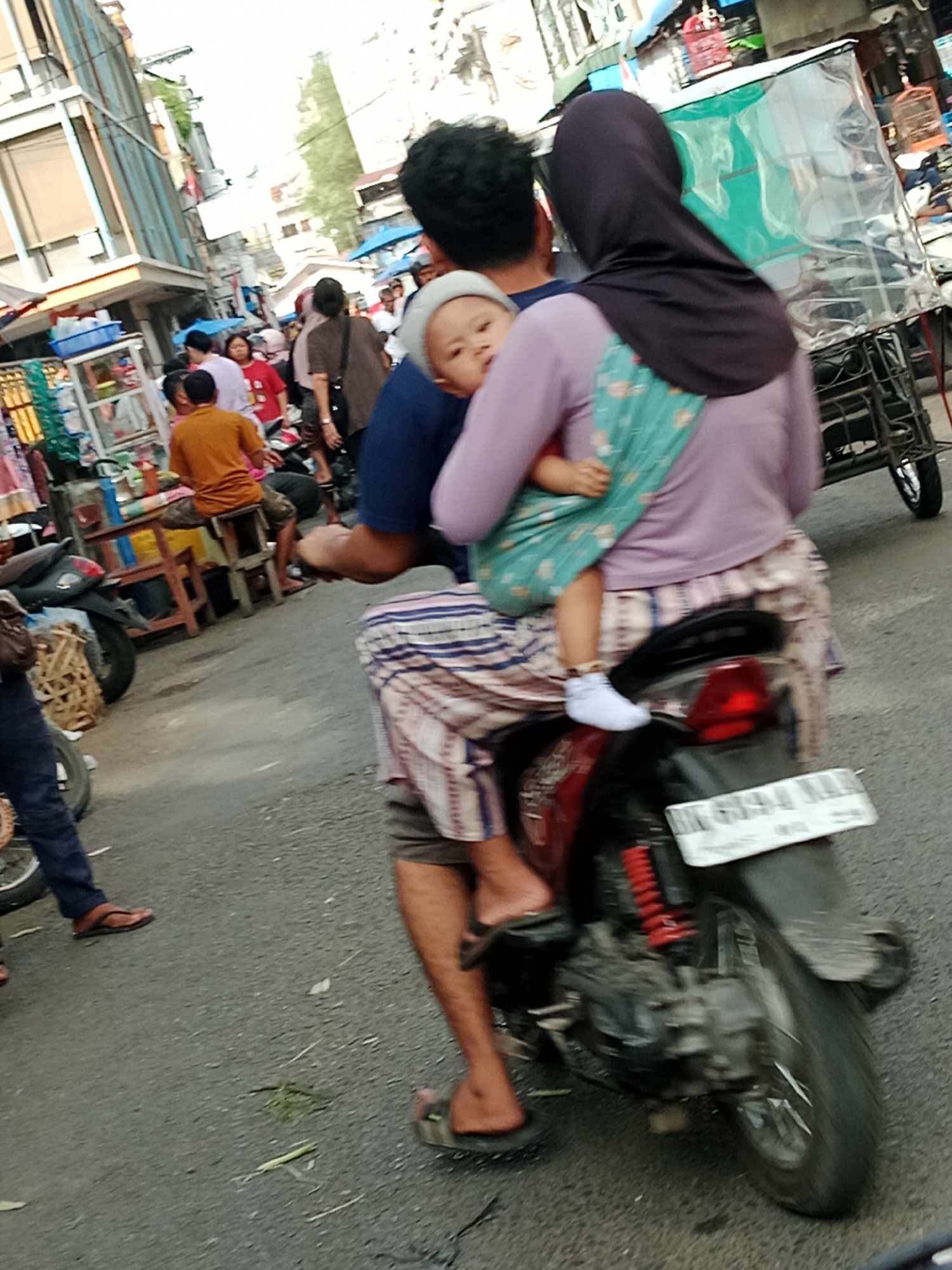 In a busy outdoor marketplace in the City of Binjai, Indonesia, a woman wearing a dark hijab and a lavender long-sleeved shirt rides on the back of a motorcycle. She is holding a baby wrapped in a light blue and patterned fabric carrier. The baby is wearing a white bonnet and white shoes. The woman's patterned skirt is visible, as is the rear of the motorcycle. The scene is crowded with other people walking and standing, and market stalls are visible in the background, suggesting a vibrant and bustling atmosphere.