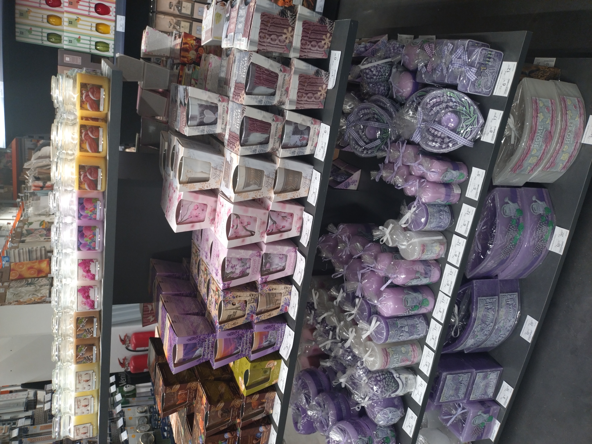 This image shows a store shelf filled with various scented candles and home fragrance products. The candles are arranged in neat rows and columns, showcasing different scents, colors, and packaging styles. On the left, there are glass jar candles with labels indicating their scents, ranging from fruity to floral. The right side of the shelf displays boxed candles and other fragrance items in shades of purple, often tied with decorative ribbons and encased in clear plastic wrapping. Price tags are visible next to each product, indicating different prices. The scene is likely taken in a retail environment, potentially a home goods store or department within a supermarket. The OCR results suggest the presence of brands or scent names like "Strawberry," "Orchid," "Angel," and "Cosy Morning." While the city of Stara Zagora, Bulgaria, is mentioned in the prompt, there is no independent information about it within the image itself, nor any information about a specific location within the city.