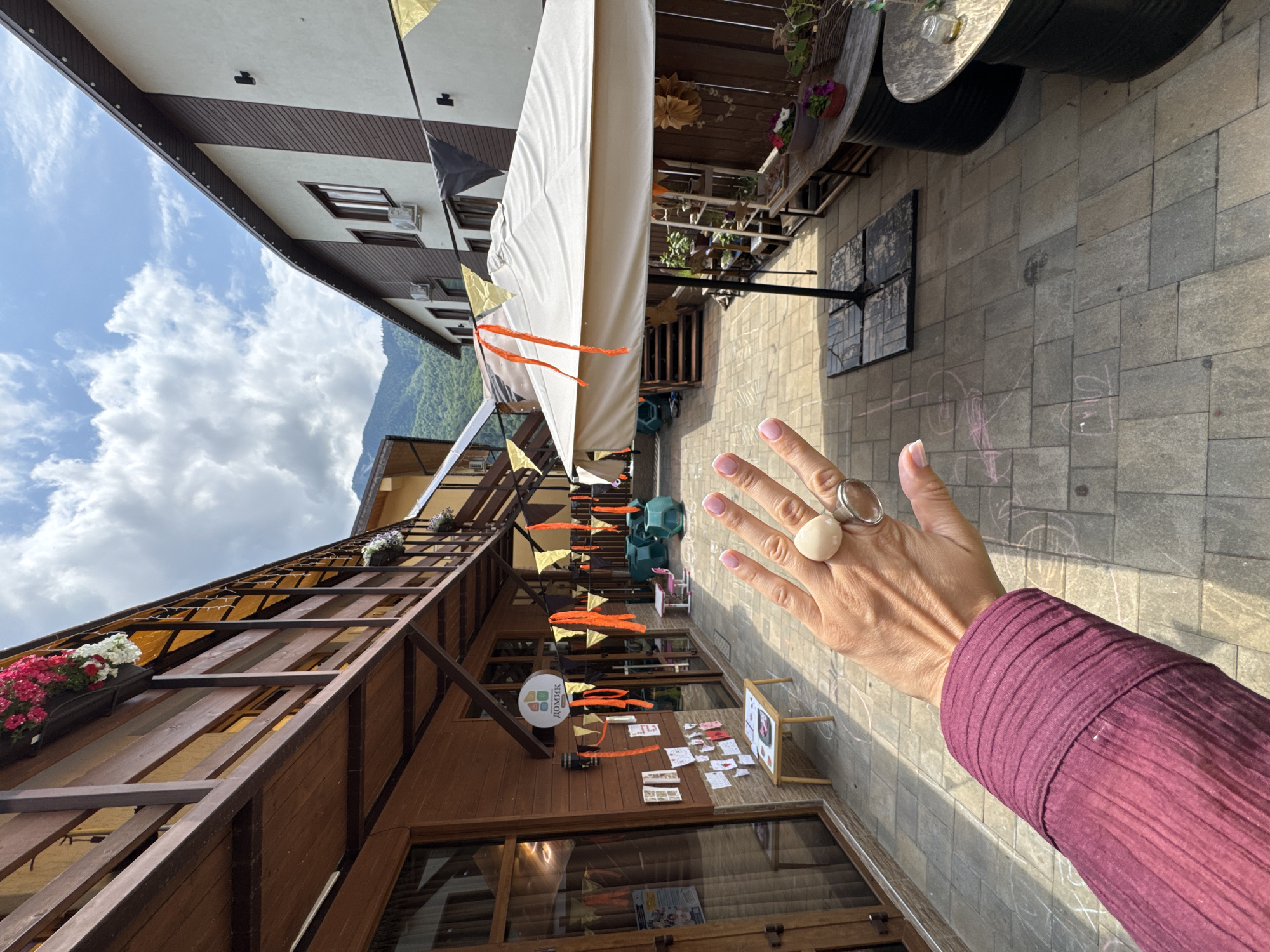 The image depicts an outdoor courtyard, likely in the city of Krasnaya Polyana, Russia, given the mountainous backdrop. In the foreground, a person's hand with manicured nails and rings is visible, extended towards the camera. The hand is positioned over a paved courtyard with chalk drawings on the ground. The courtyard is lined with wooden buildings featuring balconies and windows. String lights and festive pennants in orange and gold are draped across the scene, suggesting a celebratory or event atmosphere. A large white umbrella stands in the middle of the courtyard, providing shade. Several tables and seating areas are scattered around, some made from repurposed barrels. Plants in pots are also visible on some of the tables. In the background, a mountain range covered in greenery is faintly visible under a partly cloudy sky.