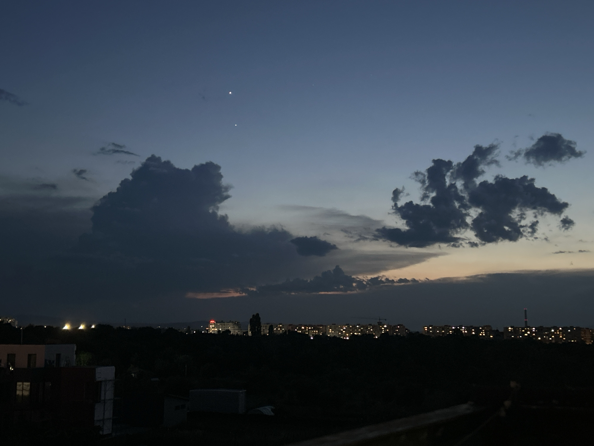 The image depicts a twilight cityscape, likely in Sofia, Bulgaria, given the context. The sky is a deep blue, transitioning to lighter hues near the horizon where a sliver of orange is visible. Two bright celestial bodies, possibly planets or stars, are prominent in the upper sky, with one slightly above and to the right of the other. Dark, dramatic clouds dominate the middle section of the sky, with one large, heavy cloud formation on the left and a more dispersed cluster on the right.
Below the sky, a silhouette of a city stretches across the frame. Numerous buildings are visible, illuminated by warm, yellow lights, suggesting a residential or commercial area. Some taller structures, possibly apartment buildings, are discernible. A dark, dense band of trees or foliage separates the foreground from the city lights. In the immediate foreground, the dark shapes of several buildings and structures can be seen, silhouetted against the fading light. The overall atmosphere is calm and serene, capturing the transition from day to night.