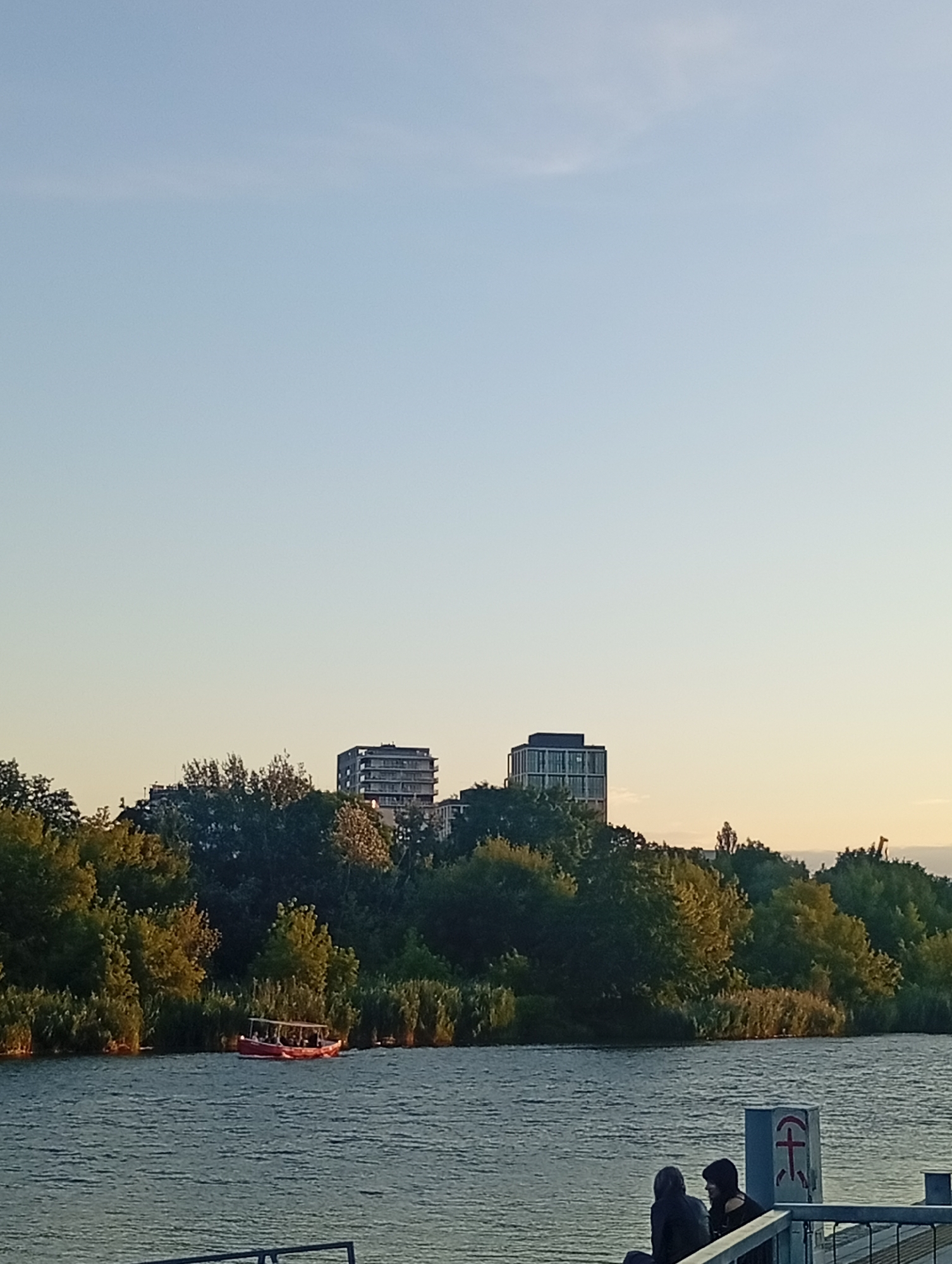 This is a medium-shot, vertical, outdoor photograph taken at a distance, likely during the golden hour, depicting a waterfront scene. The sky occupies the upper two-thirds of the frame, exhibiting a clear, pale blue hue with a hint of yellowing near the horizon. Below the sky, a dense line of lush green trees and vegetation forms a natural border along the far bank of a body of water.
Peeking above the trees, two modern, multi-story buildings are visible. The building on the left appears to be slightly shorter and darker, while the building on the right is taller with a lighter facade and visible balconies. A single, tall, thin tree stands in the middle distance, to the right of the taller building. Further to the right and further in the distance, a faint outline of what may be a crane or tower is visible against the sky.
In the foreground, the calm surface of a body of water reflects the warm light. On the water, a small, red, open-topped boat carrying several passengers is positioned near the tree line. On the right side of the frame, in the immediate foreground, two individuals are seated on a pier or walkway. They are partially obscured and facing away from the camera. A dark grey structure with a red symbol resembling a stylized anchor or cross is attached to a pillar in front of them. A metal railing runs along the edge of the walkway in the lower portion of the image.
The overall impression is one of a peaceful, urban waterfront setting at dusk, with a blend of natural scenery and modern architecture. The specific location is the Wrocław Water Sports Center (Wrocławskie Centrum Sportów Wodnych) on the Oder River (Odra River) in Wrocław, Poland, given the distinctive architectural style of the buildings and the presence of water sports facilities.