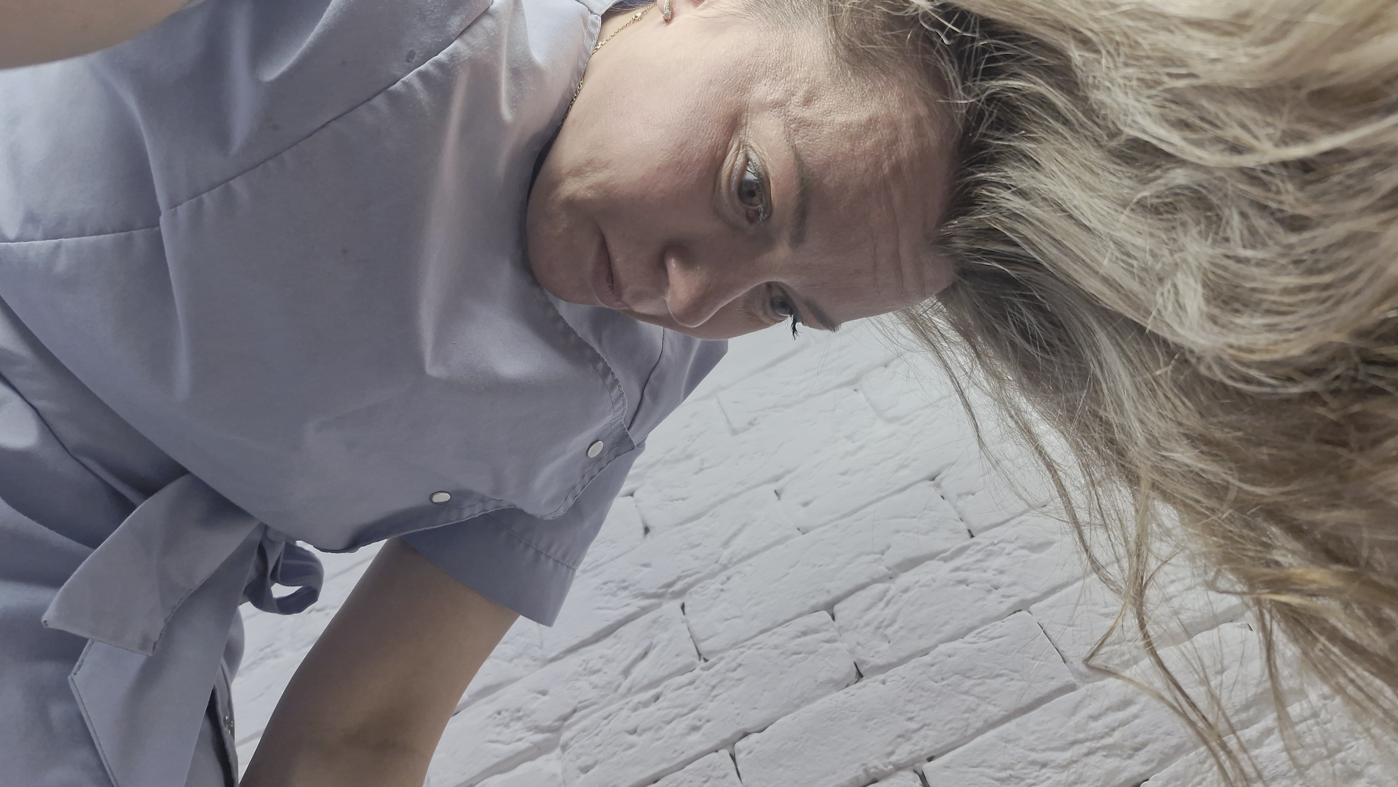 An eye-level shot captures a woman with blonde hair, dressed in a light blue professional uniform, looking downwards with a focused expression. The camera is positioned below her, angled upwards, suggesting she is leaning over an object or person out of frame. Her long, light-colored hair falls loosely around her face and across the right side of the image. She has fair skin, light-colored eyes, and visible fine lines on her forehead and around her eyes. A small gold earring is visible on her left ear.
She is wearing a light blue tunic or smock-style top, featuring snap buttons and a tie-waist detail on the left side, indicative of professional attire. The background is a textured white brick wall, suggesting an indoor professional environment such as a clinic, salon, or workspace. Her attentive gaze implies she is engaged in a task requiring concentration. No text is visible in the image. The scene is set in Otchor, Russia.