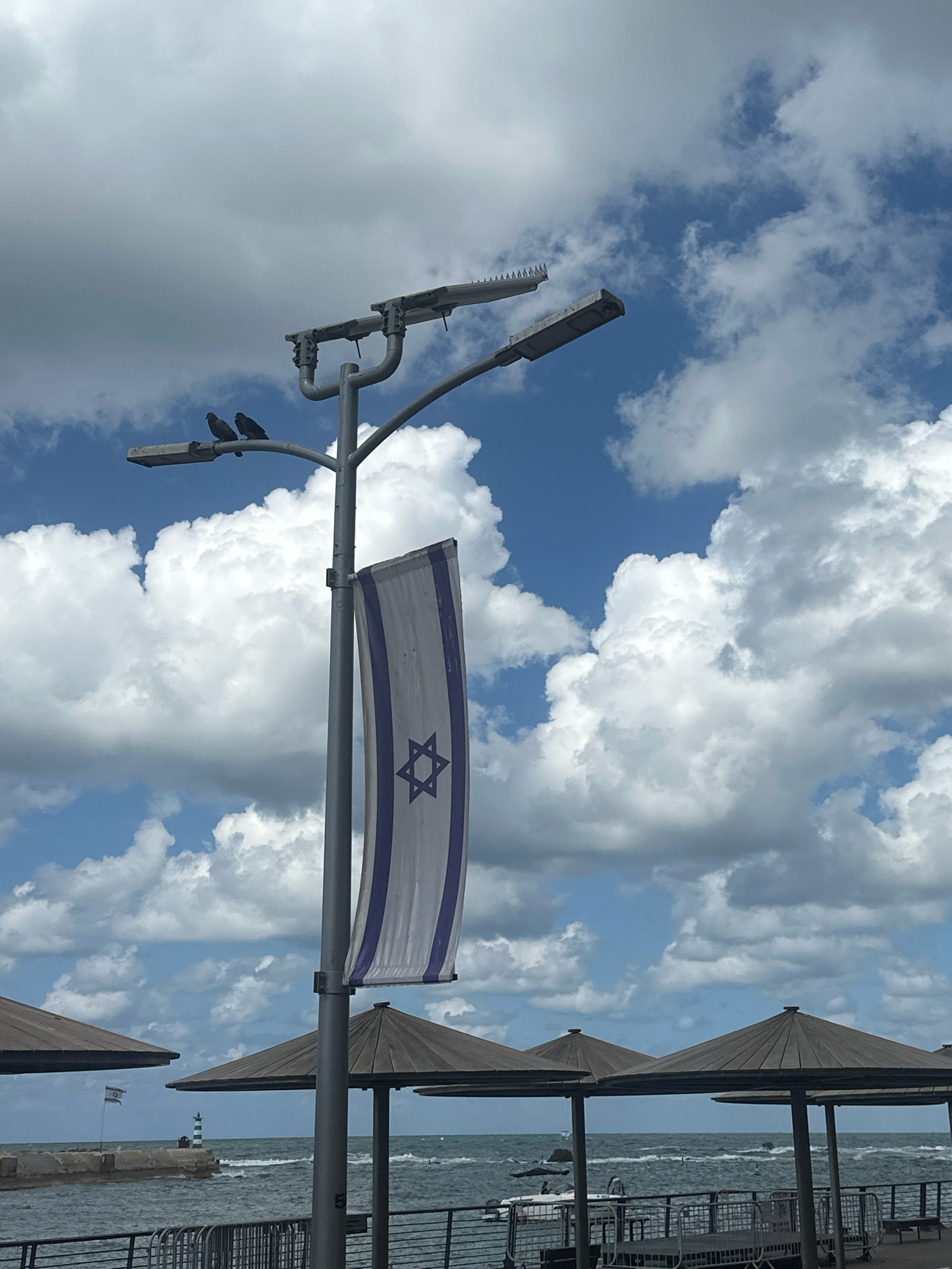 This image captures a scene at the Tel Aviv port area, with the Mediterranean Sea in the background under a cloudy sky. A lamppost prominently features a hanging Israeli flag, its Star of David visible. Two birds are perched on one of the lampposts. In the distance, a lighthouse stands on a breakwater, and several umbrella-like structures provide shade along the waterfront. Boats are visible in the water. The overall ambiance suggests a day at the beach or port.