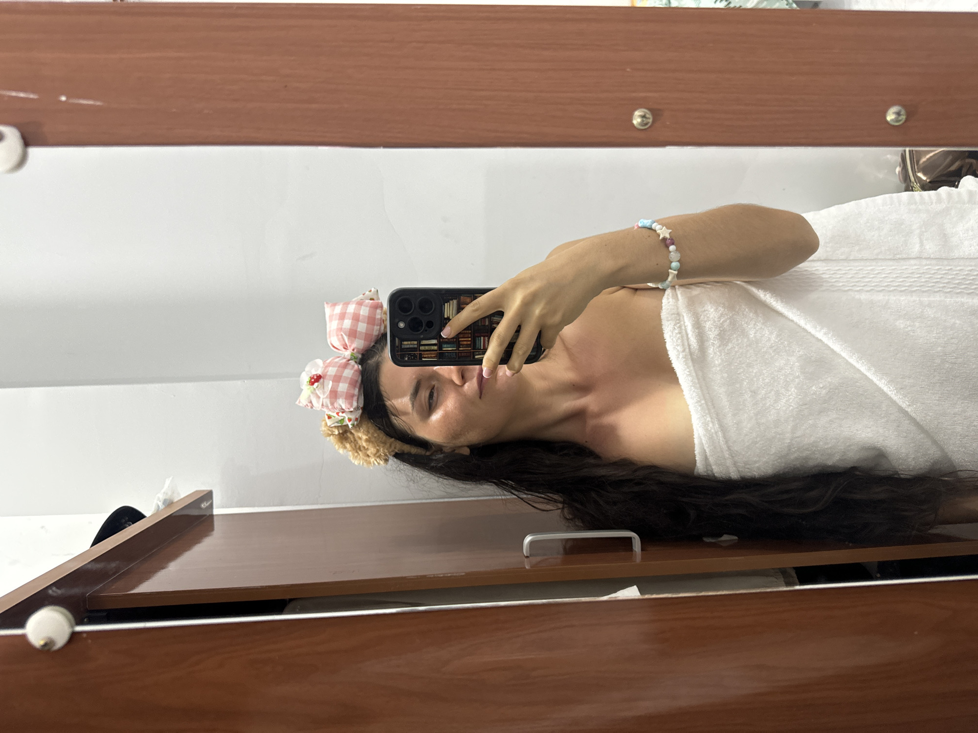 A woman is taking a selfie in a mirror. She is wearing a white towel and a pink checkered headband with flowers. She is holding a black smartphone in her right hand. The mirror is framed in wood. The scene appears to be indoors, possibly in a bathroom or spa. The location is Mũi Né, Vietnam.
