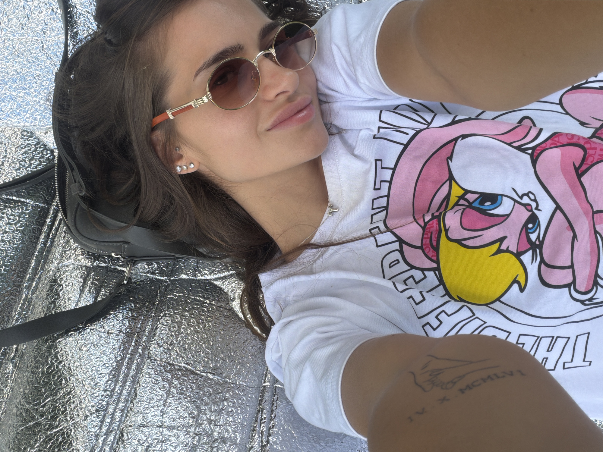 A woman is lying down, taking a selfie. She has long brown hair and is wearing round sunglasses with orange temples and a white t-shirt with a cartoon graphic. She has multiple piercings in her left ear and a tattoo on her upper left arm. She is lying on a shiny silver surface with a black bag next to her. The location is Unknown, Unknown.