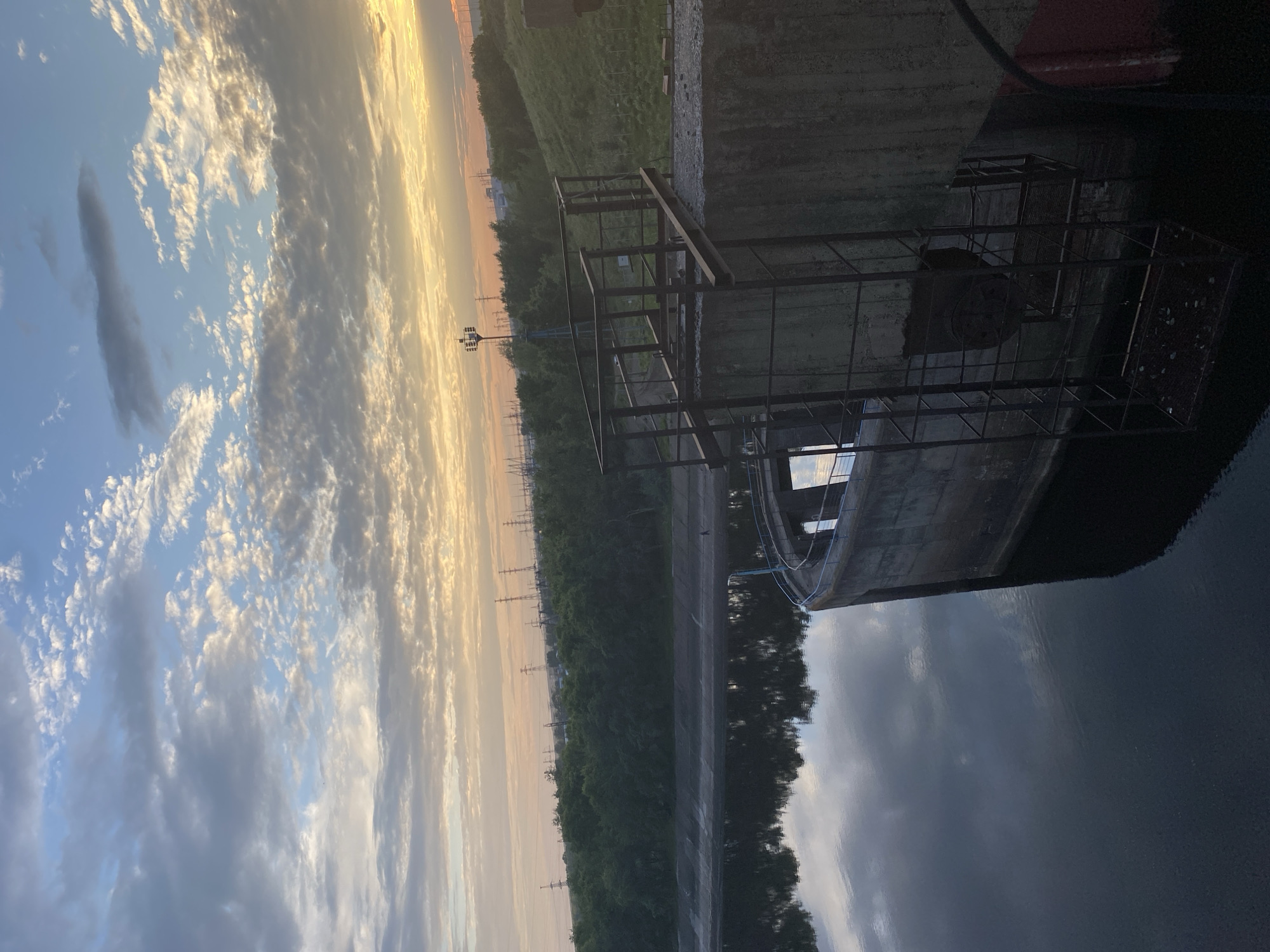 The image shows a wide shot of a dam and reservoir at sunrise. The sky is filled with dramatic clouds, illuminated by the warm tones of the rising sun. The dam structure is made of concrete and has a metal ladder attached to its side, leading down to the water. The reservoir is calm and reflects the sky and surrounding greenery. In the background, there are trees and power lines, suggesting an industrial or semi-urban setting. A single person is visible walking on the bank of the reservoir. The scene appears to be tranquil and serene, despite the industrial elements in the background. The location is the Votkinsk Dam in Chaikovsky, Russia.