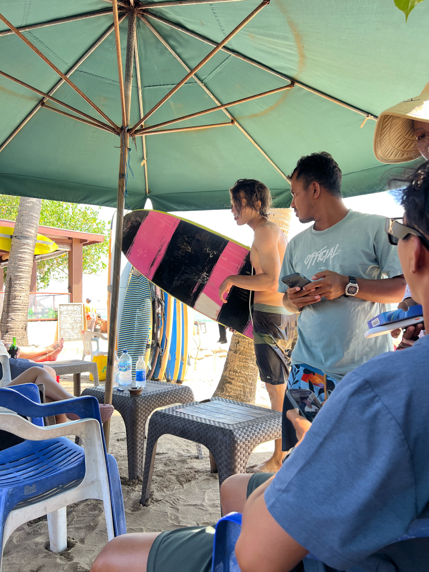 Under a large green umbrella at a beach in Kuta, Indonesia, a group of people are gathered. A young man, shirtless and wearing patterned shorts, holds a pink and black surfboard. To his right, another man in a light blue t-shirt with the word "OFFICIAL" printed on it, and colorful swim shorts, looks down at his phone. A third person, partially visible in the foreground, is looking at their phone, and a fourth person wearing a straw hat is also in view. In the background, several surfboards are propped up, and a building with a red roof is visible. Two small tables are placed on the sandy beach, one with two bottles of water and an ashtray, and the other empty. The scene suggests a casual, outdoor gathering, possibly before or after a surfing session, at a beachside location.