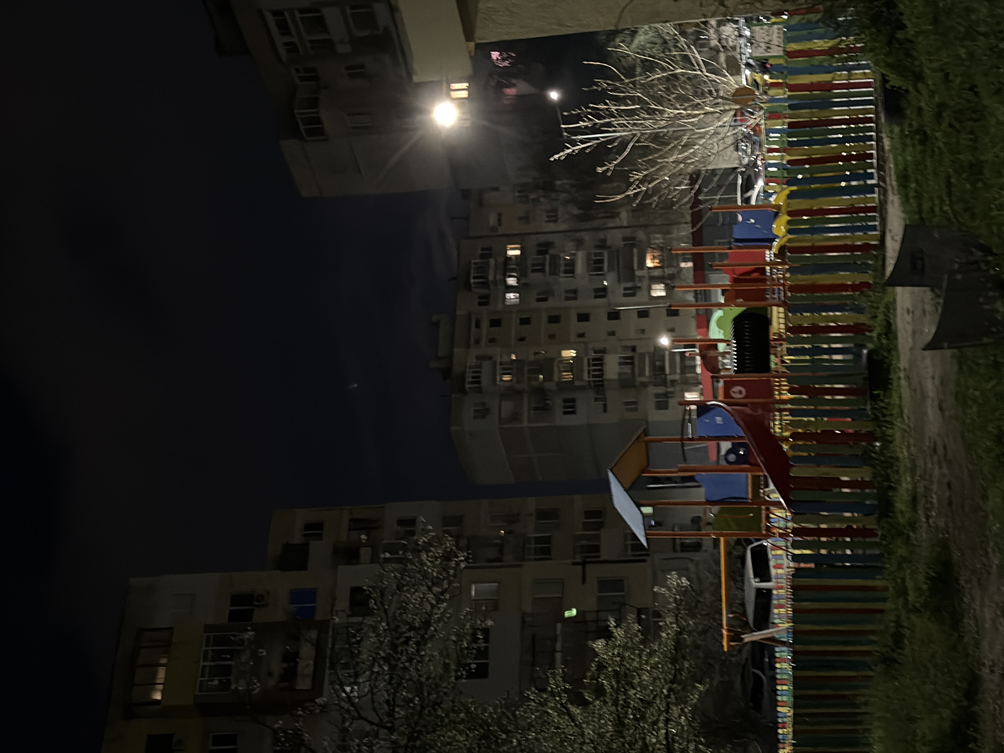 The image shows an outdoor scene in Varna, Bulgaria, at night. Several apartment buildings surround a brightly colored playground. The buildings appear to be typical residential structures, with numerous windows. A street lamp illuminates the area near the playground. There are trees in the foreground and along the right side of the image.