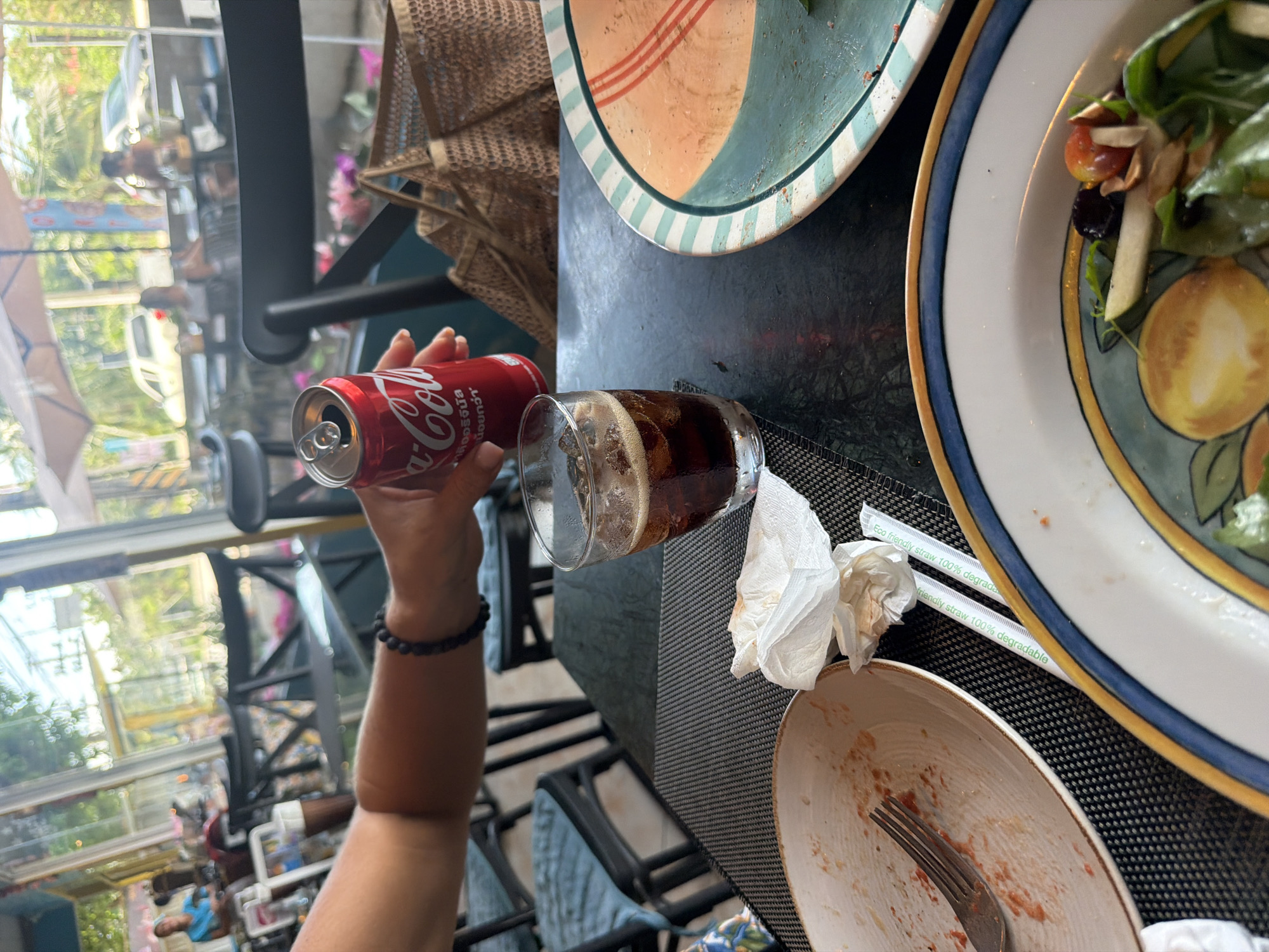 An outdoor cafe scene is captured, likely in Baan Bo Phut, Thailand, with a glass of Coca-Cola on a dark, textured table. A person's arm, adorned with a black beaded bracelet, holds a partially consumed can of Coca-Cola. The table is also set with a white paper napkin, a fork on a dirty plate, and two paper straws. In the background, other diners are visible through large windows, with tables and chairs set up both inside and outside under umbrellas. Palm trees and glimpses of vehicles suggest a tropical, possibly roadside, setting.
