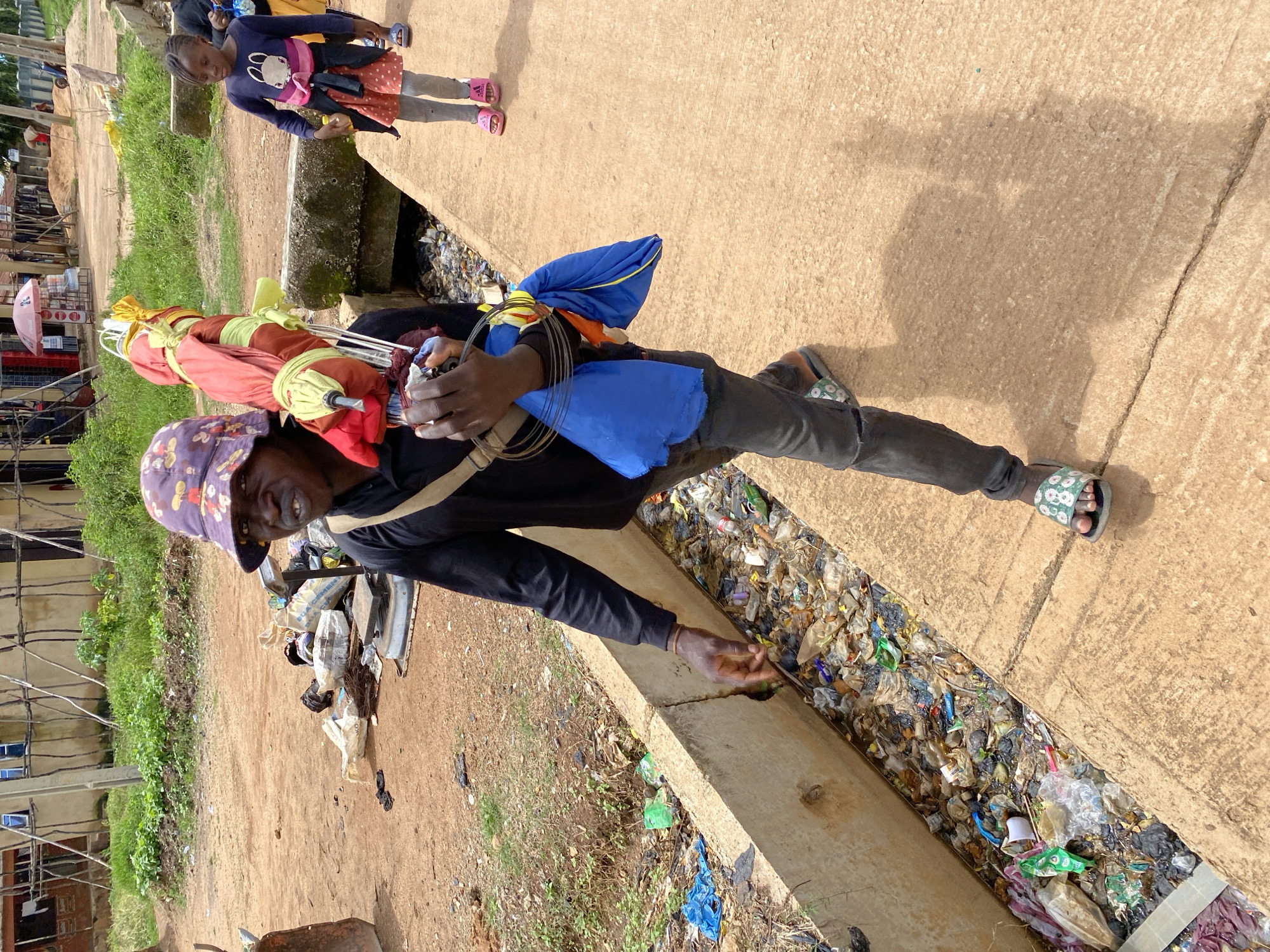 A man wearing a patterned bucket hat, a black long-sleeved shirt, and dark pants walks along a concrete walkway beside a trash-filled drainage ditch in Bukuru, Nigeria. He carries a bundle of colorful items wrapped in fabric and plastic over his shoulder, with some blue fabric tied around his arm. A young girl in a pink and orange dress and gray leggings walks behind him, also on the concrete path. The environment appears to be an urban area with unpaved ground, some greenery, and buildings in the background. The drainage ditch is heavily contaminated with various types of refuse.
