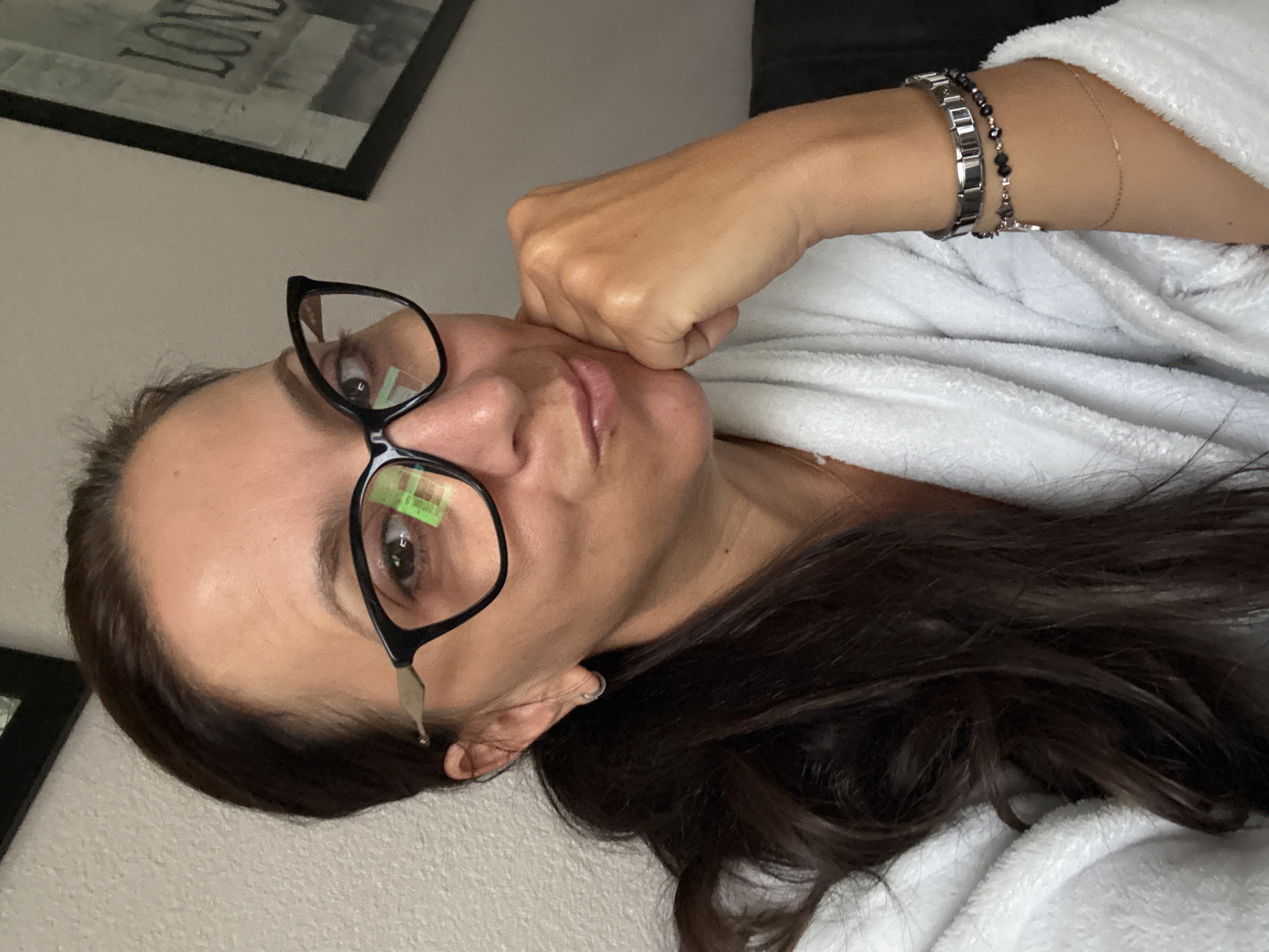 A woman with dark hair pulled back in a ponytail and wearing black cat-eye glasses is captured in a close-up, shoulder-up shot. She is dressed in a white fluffy robe and is resting her chin on her right hand. She has a delicate earring in her left ear and is wearing two bracelets on her right wrist. The background features a textured light-colored wall and a framed picture with "LONDON" visible. The image appears to be taken indoors, possibly in a home environment in Marum, Netherlands.