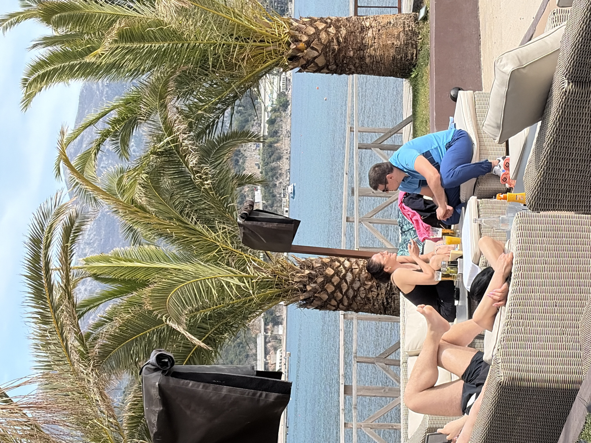 The image depicts an outdoor leisure setting, likely a resort or beach club in Budva, Montenegro, with the Adriatic Sea and coastal mountains in the background. Tall palm trees frame the scene. In the foreground, people are relaxing on wicker lounge chairs. One woman, wearing a black swimsuit, reclines on a chair. Another individual in black shorts is visible partially, reclining on the same chair. A man wearing a blue athletic shirt and dark pants is kneeling on a chair, near some beach items. Drinks and sunscreen are on a table. A wooden railing separates the lounge area from the water. A large black umbrella is also visible. The scene suggests a sunny day at a relaxing seaside location.