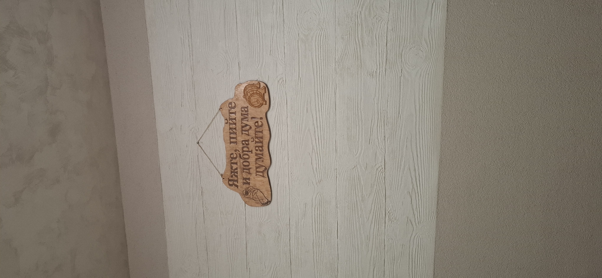 The image shows a wooden sign with Russian text and a drawing of a wine barrel and a glass, hanging on a white wooden paneled wall. The text on the sign reads "Яжте, пийте и добра дума думайте!", which translates to "Eat, drink, and think good thoughts!". The sign is suspended by a thin rope. The wall appears to be indoors, possibly in a home or a restaurant. The ceiling above the wall is textured and light brown. The bottom part of the image shows a grey textured surface, possibly another wall or a piece of furniture. There are no people or actions depicted in the image. The location in Pleven, Bulgaria is not discernible from the image.