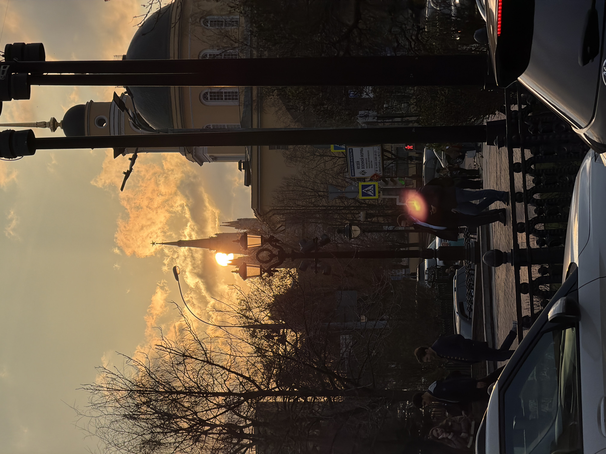 This image captures an urban street scene in Moscow, Russia, during what appears to be a vibrant sunset or sunrise. The low-lying sun casts a warm, golden light across the sky, illuminating scattered clouds and creating dramatic silhouettes of buildings and trees.
In the background, a light-colored building with a large dark dome and smaller cupolas, featuring arched windows, stands prominently. Behind it and to the left, a tall, dark spire topped with a star is silhouetted directly against the sun, suggesting a Kremlin tower. Bare deciduous trees, indicative of late autumn, winter, or early spring, frame the scene from the foreground and midground, their branches stark against the bright sky.
The street is lined with several ornate black streetlights, one of which directly intersects with the sun, causing a significant lens flare. Traffic signs, including yellow and blue pedestrian crossing symbols, are visible. On the right side of the frame, parts of several cars are present; a dark vehicle in the upper right shows illuminated red taillights, while a lighter-colored car is seen in the lower foreground.
Several pedestrians are walking on the sidewalk, their figures largely in shadow due to the backlighting. One individual in the midground appears to be holding a reflective object, creating a strong, star-like lens flare.
Visible Cyrillic text on a sign on the left clearly states "МИНИСТЕРСТВО КУЛЬТУРЫ РФ" (Ministry of Culture of the Russian Federation) and advertises the "МУЗЕЙ ВОЕННОЙ ФОРМЫ" (Museum of Military Uniforms), providing its phone number and address: "Б. НИКИТСКАЯ ул., д. 46/17, с.1" (Bolshaya Nikitskaya Street, building 46/17, structure 1), confirming the location in Moscow. The overall mood is serene yet active, bathed in the dramatic light of the rising or setting sun.