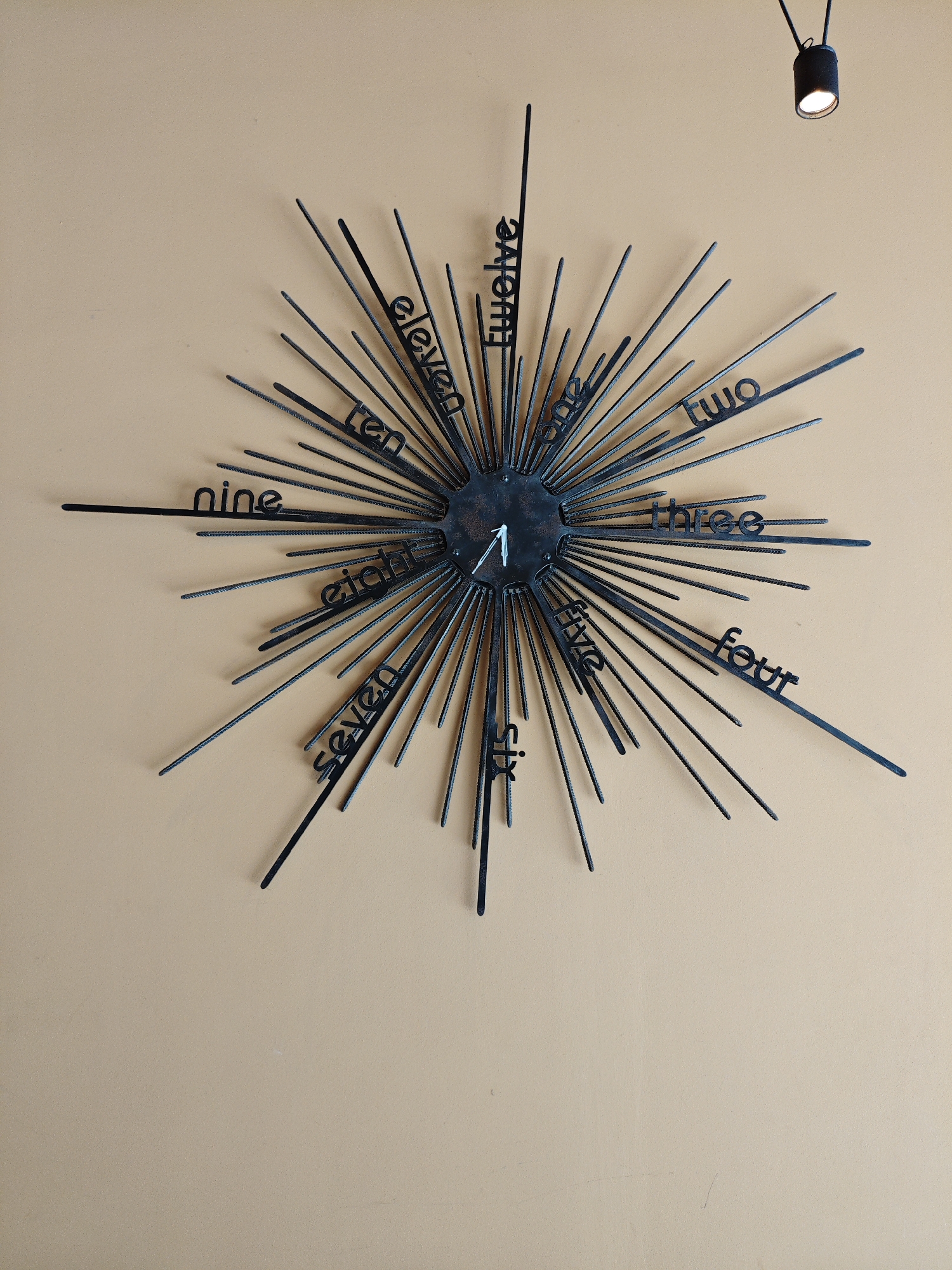 A starburst-shaped metal clock is mounted on a beige wall. The clock features numbers one through twelve written out in black, stylized text. A small spotlight is visible in the upper right corner of the image. The clock is centrally located in the frame, with its spiky spokes extending outwards. The wall appears to be a flat, painted surface. There are no people visible in the image. The image does not provide any information about the city of Alushta, Ukraine, or any context related to that location.