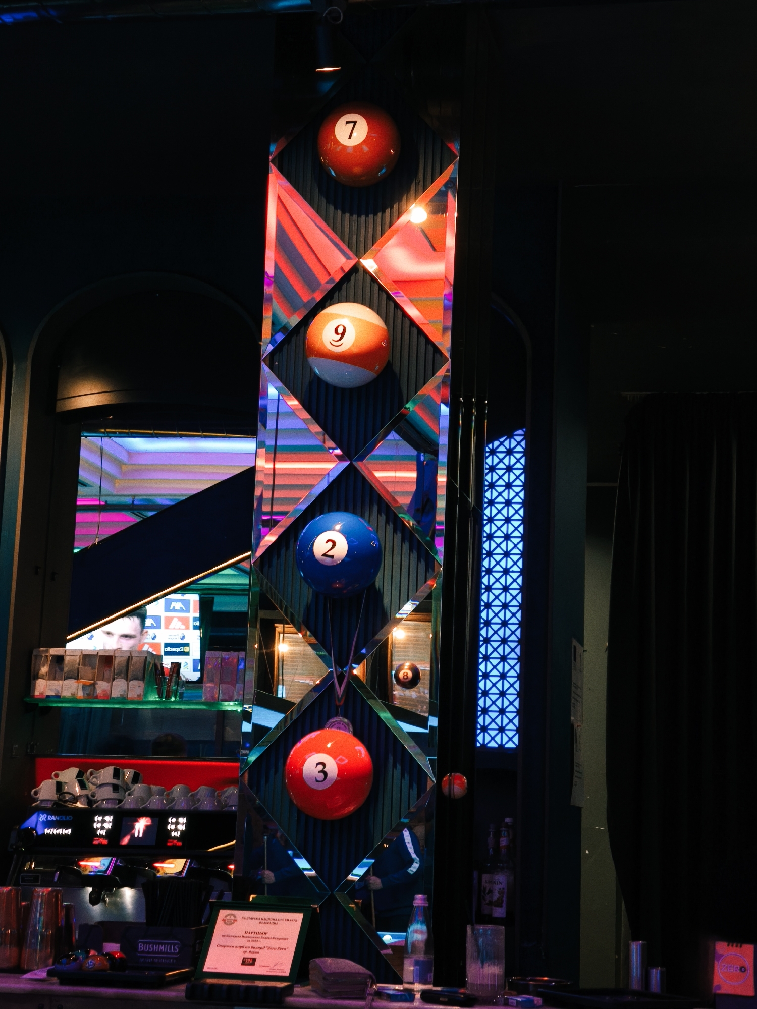 The image captures a vertical decorative element in what appears to be a bar or entertainment venue, possibly in Varna, Bulgaria. The element features a mirrored structure with diamond-shaped panels, each holding a billiards ball with different numbers: 7 (red), 9 (orange and white striped), 2 (blue), and 3 (red). An 8-ball is reflected in one of the mirror panels.
Below the decorative element, there is a bar area with a coffee machine (RANCILIO), stacked coffee cups, bottles of BUSHMILLS Irish Whiskey, and a sign in Bulgarian, indicating a sponsorship or partnership with the "Zero Fern" billiards club. A television screen showing a person's face and what appears to be a sporting event, is reflected in the mirror.
A mirrored wall with blue neon cross design can be seen behind the billiard balls. The scene is illuminated with colorful lighting, creating a lively and vibrant atmosphere. A dark curtain is on the right side.