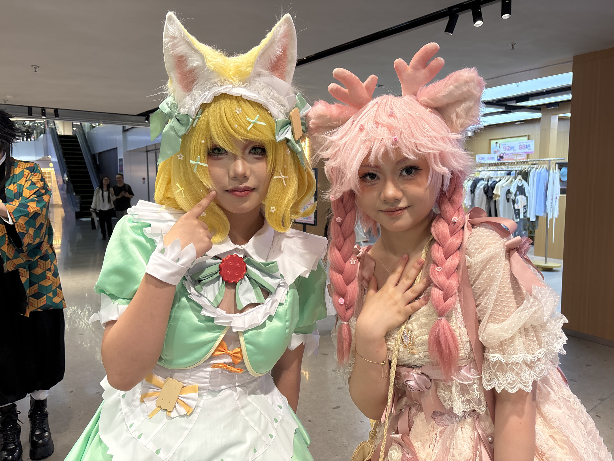 Two young women in elaborate cosplay costumes stand close together indoors. The woman on the left is wearing a yellow wig with large animal ears, a white bonnet, a light green and white dress with ribbons and bows. She is touching her face with her index finger. The woman on the right has pink hair styled in braids with a pink, antler-like headpiece. She is wearing a pink and white lace dress with ribbons. Her left hand is resting near her chest.
In the background, slightly out of focus, is an escalator and several people. Behind the women is a clothing rack with various garments. The setting appears to be a retail space, potentially in the city of Liangjiang, China.