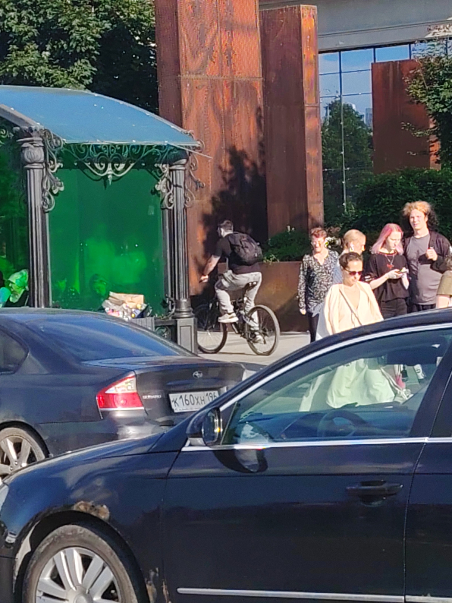 In Yekaterinburg, Russia, on a sunny day, a man rides a BMX bike. He's wearing a black t-shirt and grey shorts, with a backpack on. He's biking past a bus stop with a green glass enclosure and ornate metalwork. Behind the bus stop, there's a modern building with large windows reflecting the sky and trees. A group of people are walking near the building. One woman has pink hair and is looking at her phone. Another woman wears sunglasses and a light-colored dress. A dark-colored Subaru is parked nearby, its license plate readable as "K160XH 196". Another dark car is in the foreground, partially obscuring the view. The scene suggests a casual day out in the city.