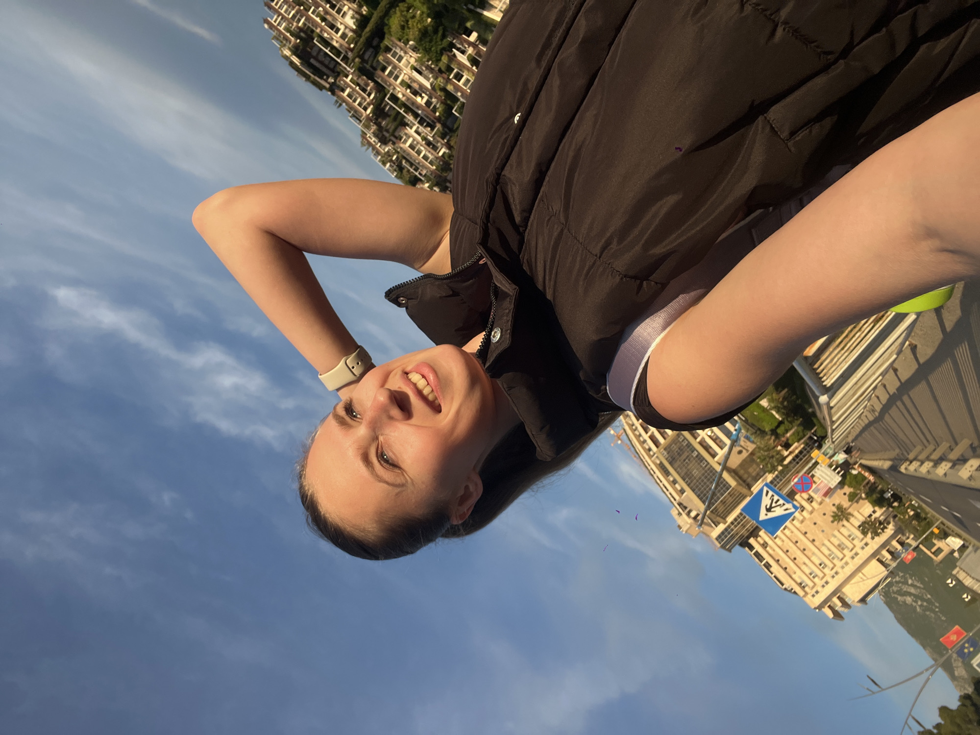 A woman is standing on a bridge in Budva, Montenegro, with the city skyline visible in the background. She is wearing a black vest and a smartwatch. The sky is blue with some clouds. Traffic signs and buildings are visible in the background.