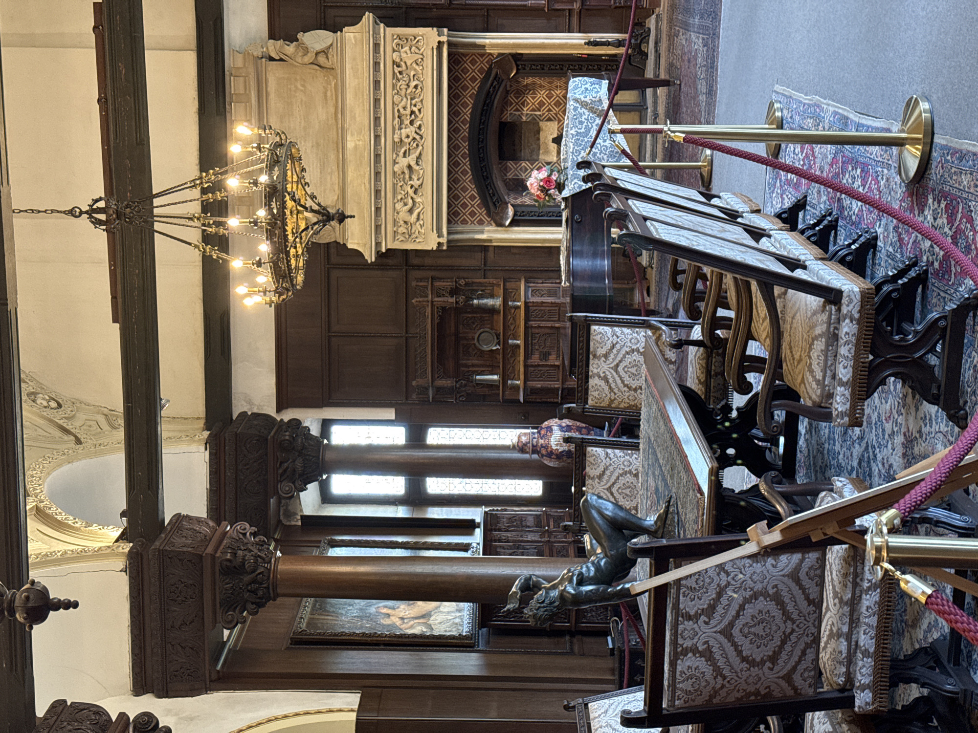 The image captures an interior scene, likely within Reichsburg Cochem, Germany. A large, ornate chandelier hangs from the ceiling, illuminating a space with dark wood paneling. A monumental carved fireplace stands on one end. In the foreground, chairs with patterned upholstery are neatly arranged behind a velvet rope. A bronze statue of a reclining man is visible on the right. A row of windows lets in natural light, partially illuminating artwork on the wall. The interior exudes a historical ambiance.