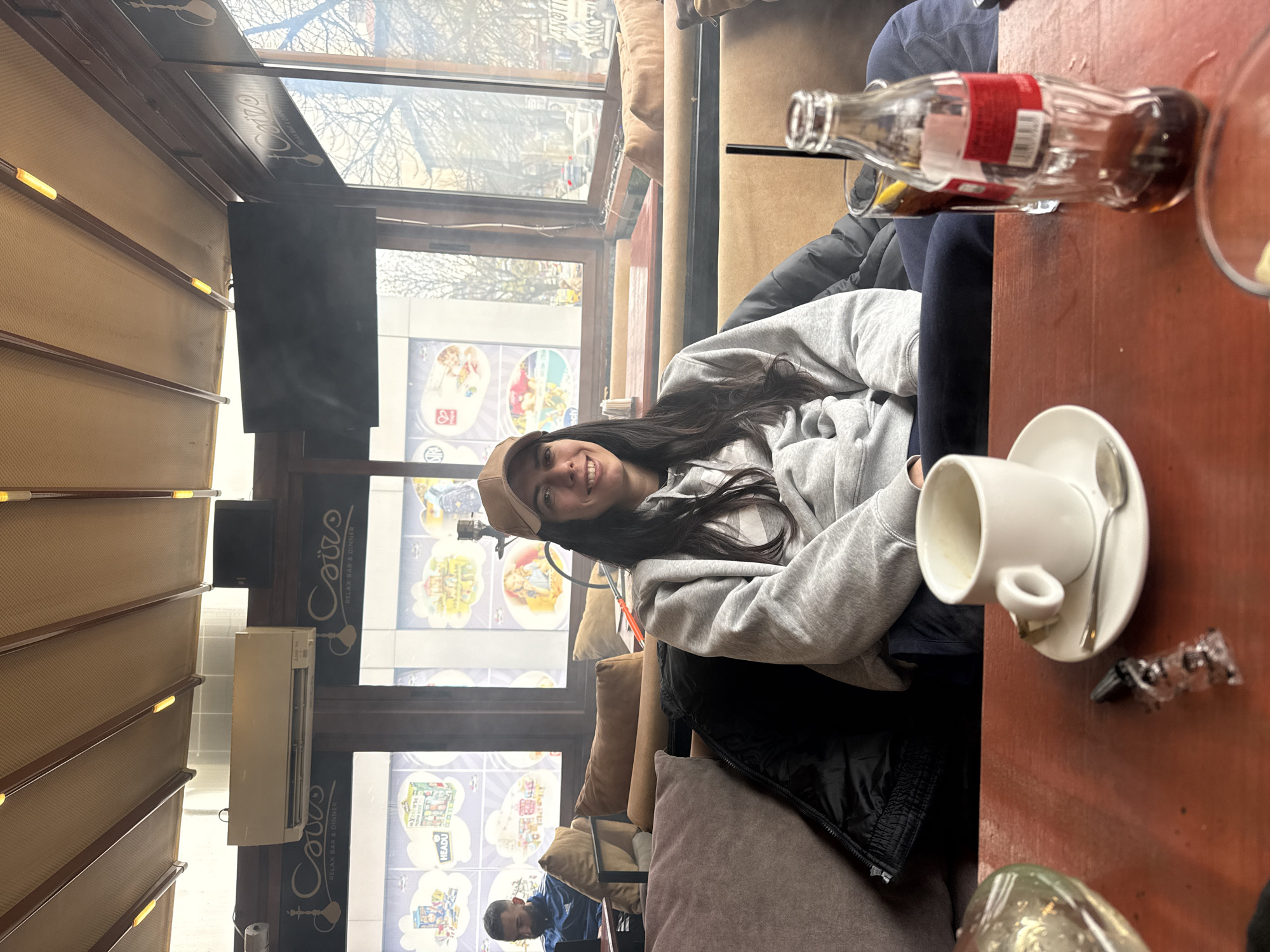 The image shows a woman seated in the "Cairo" Relax Bar & Dinner in Sofia, Bulgaria. She is wearing a gray hoodie and a brown cap, and she is smiling at the camera. Next to her on the wooden table is a half-empty Coca-Cola bottle, a white ceramic coffee cup and saucer with a spoon, and some cellophane packaging remnants. In the background, there are windows with decorative images and a view of the outside. Another person in a blue shirt is seated in the distance. Interior decor is a beige color and lighting is warm.