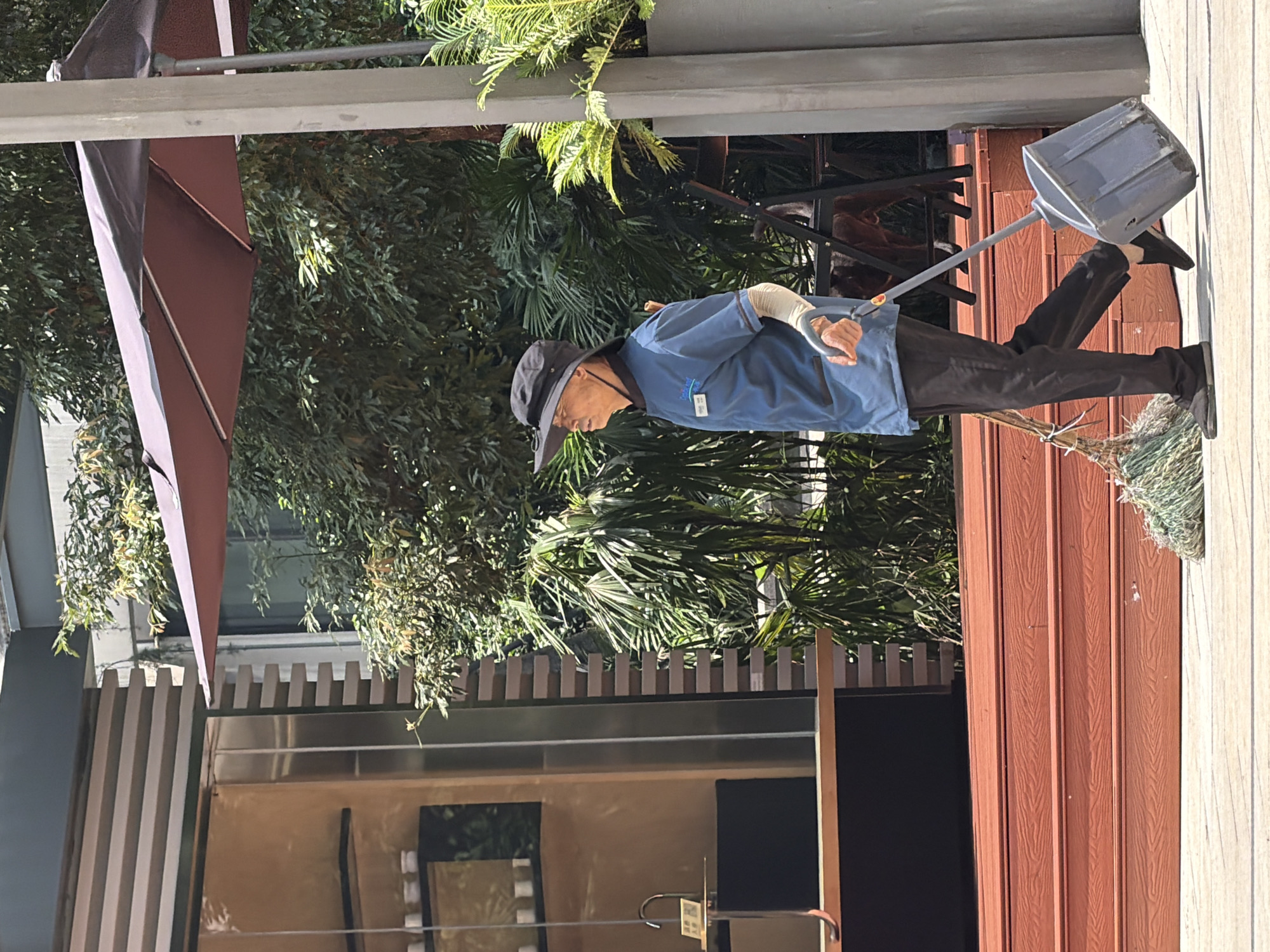 An elderly Asian man wearing a wide-brimmed hat, a blue uniform shirt with a name tag, and dark pants, walks to the right while pulling a dustpan and broom behind him on a paved surface. He has on light-colored arm warmers. Behind him are red wooden steps, lush green trees, and a building with a glass storefront. A large gray pole is to the right of the man. The scene appears to be outdoors in a public area, possibly a plaza or walkway adjacent to a commercial establishment in Yuzhong District, China.
