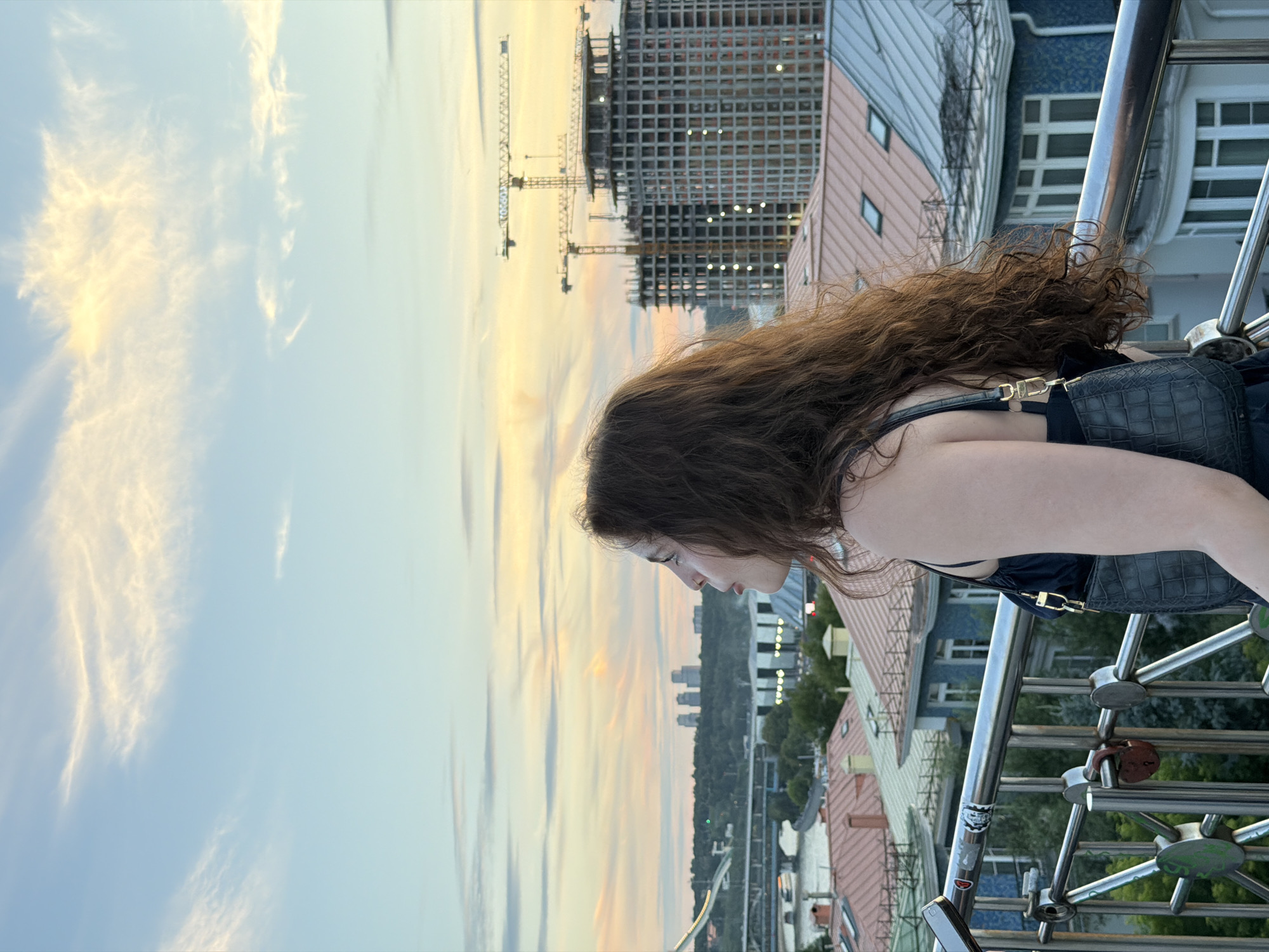 A woman with long, curly brown hair stands on a railing, looking out at a city skyline during sunset. She is wearing a dark dress and carrying a black, textured handbag. The background shows a river, a bridge, and several buildings, including some under construction with cranes. The sky is filled with soft clouds in shades of blue, pink, and yellow. The scene is likely from a high vantage point, possibly a bridge or rooftop, in Moscow, Russia, given the architectural style and presence of construction.