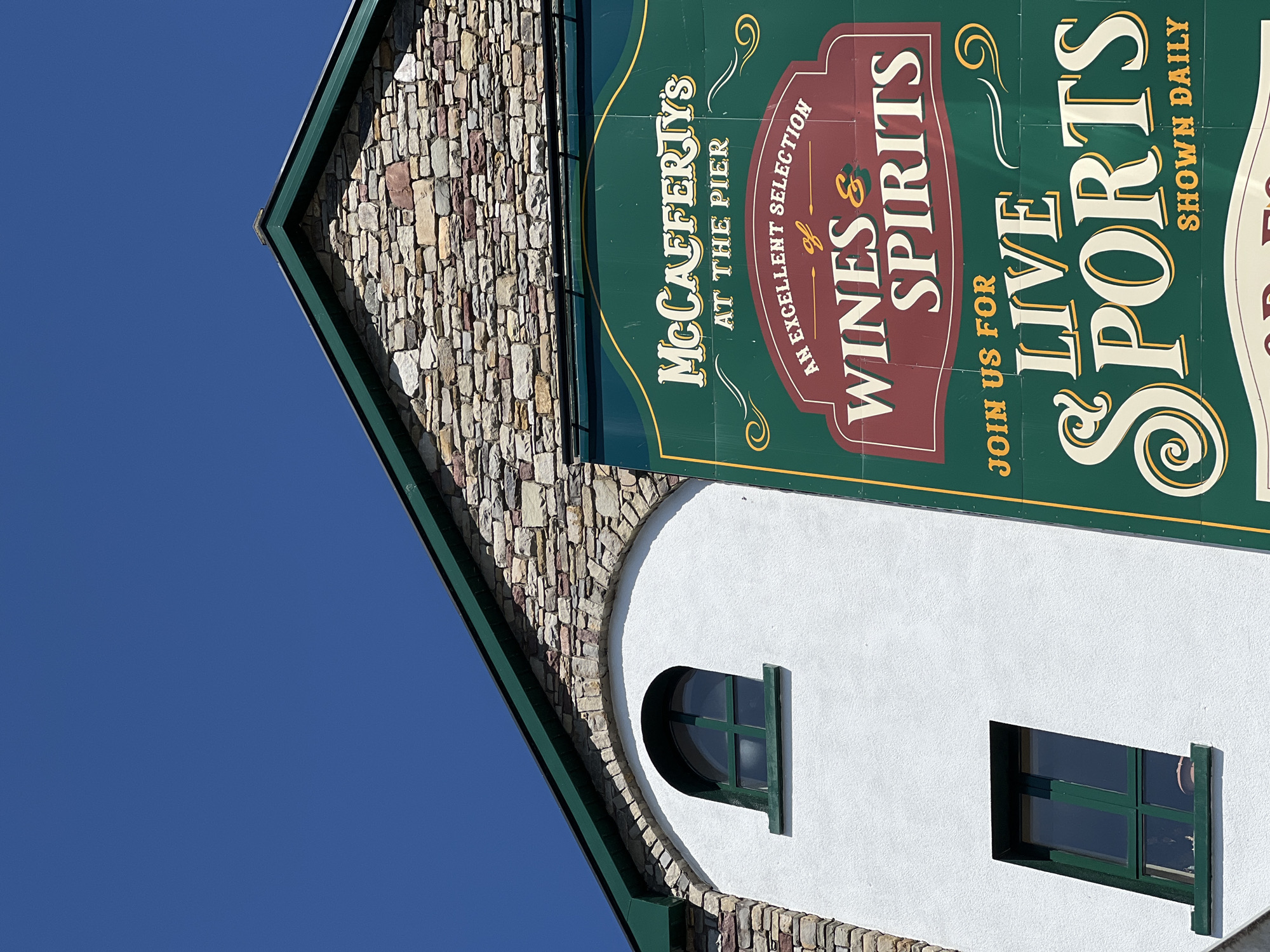 The image shows a close-up of the exterior of a building with a stone-clad upper section and a white plastered lower section. A large green and maroon sign is attached to the front of the building. The sign features text that reads "McCafferty's AT THE PIER AN EXCELLENT SELECTION of WINES & SPIRITS JOIN US FOR LIVE SPORTS SHOWN DAILY". There are two green-framed windows visible on the white plastered section of the building, one arched and one rectangular. The sky above is clear blue. The scene is located in Donegal, Ireland.