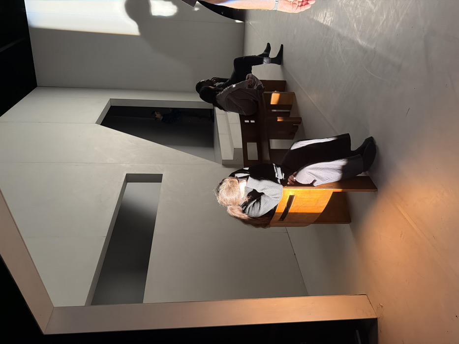 This image depicts an indoor, minimalist setting, likely a studio or gallery in Paris, France. The environment features stark white walls and a light-colored floor, accented by geometric architectural cutouts and an angled doorway in the background. Overhead, a dark ceiling is visible. The scene is illuminated by strong, directional light sources, casting sharp shadows and creating a prominent orange light spill on the floor in the foreground left.
In the foreground left, a person with long brown hair, wearing a dark ribbed long-sleeved top and dark lower garments, is seated on a minimalist wooden chair. Their head is bowed and obscured by their arm, suggesting a posture of rest or distress.
In the mid-ground right, two individuals are seated on a minimalist wooden bench. The person closer to the foreground has dark hair and wears a light-colored top, facing towards the right. Next to them, another person with dark hair, wearing a dark top and dark pants, displays light-colored socks, also facing right. They appear to be observing or engaged in a quiet interaction.
In the background, within a dark, angular doorway opening on the right side of the main wall structure, a partially obscured figure is standing, appearing to observe the scene. A partial view of a hand and arm, possibly belonging to a fourth person or crew member, is visible on the far right edge of the frame. The overall atmosphere is quiet and contemplative, characterized by the stark setting and dramatic lighting.