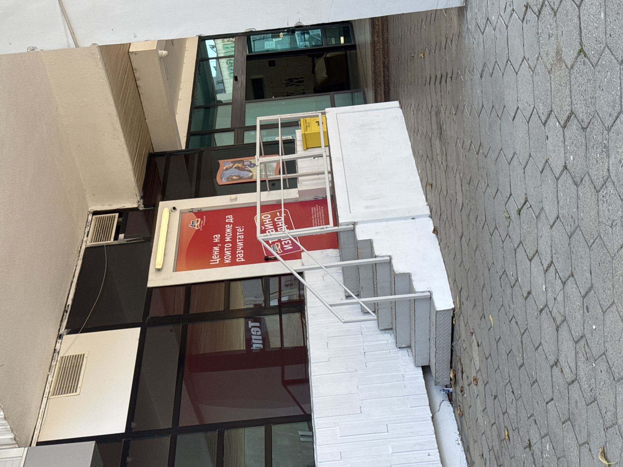 The image shows the exterior of a building with a white facade and dark glass windows. A set of gray concrete stairs with a white metal handrail leads up to a red door. Above the door, a sign reads "dm" and below it, in Bulgarian, "Цени, на които може да разчитате!" which translates to "Prices you can count on!". To the left of the stairs, a white textured wall is visible. A yellow mailbox is positioned near the top of the stairs. The ground in front of the building is paved with hexagonal gray concrete blocks. In the background, through the glass doors, a dimly lit interior with furniture is discernible. The overall scene appears to be an entrance to a commercial establishment in Kazanlak, Bulgaria.