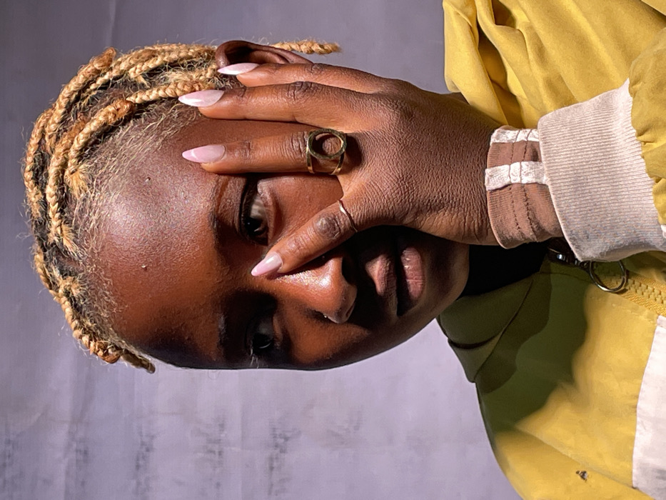 A close-up portrait features an individual with dark skin, positioned slightly off-center, looking towards the viewer's left. The person has light blonde, braided hair styled in short twists or locs. A small piercing is visible on the left nostril. Their right hand is pressed against the right side of their face, partially covering the cheek and temple. The fingers display long, almond-shaped nails with a light pink or nude polish. Two gold-colored rings adorn the right hand: a thin band on the ring finger and a larger ring with an open circular design on the middle finger. The individual wears a yellow jacket with a prominent zipper, featuring a metallic pull ring. A white horizontal stripe is visible on the lower left portion of the jacket. The jacket's cuff is a light, ribbed fabric, underneath which a brown or tan sleeve/wristband is visible. The background is a plain, light-colored surface with subtle vertical creasing, suggesting a studio backdrop or fabric wall. Strong, directional lighting illuminates the subject from the right, creating highlights on the skin and casting soft shadows on the left side of the face and behind the hand. The image does not contain any discernible visual information to indicate the specific city of Barakin Akawo, Nigeria, or any other geographical location.